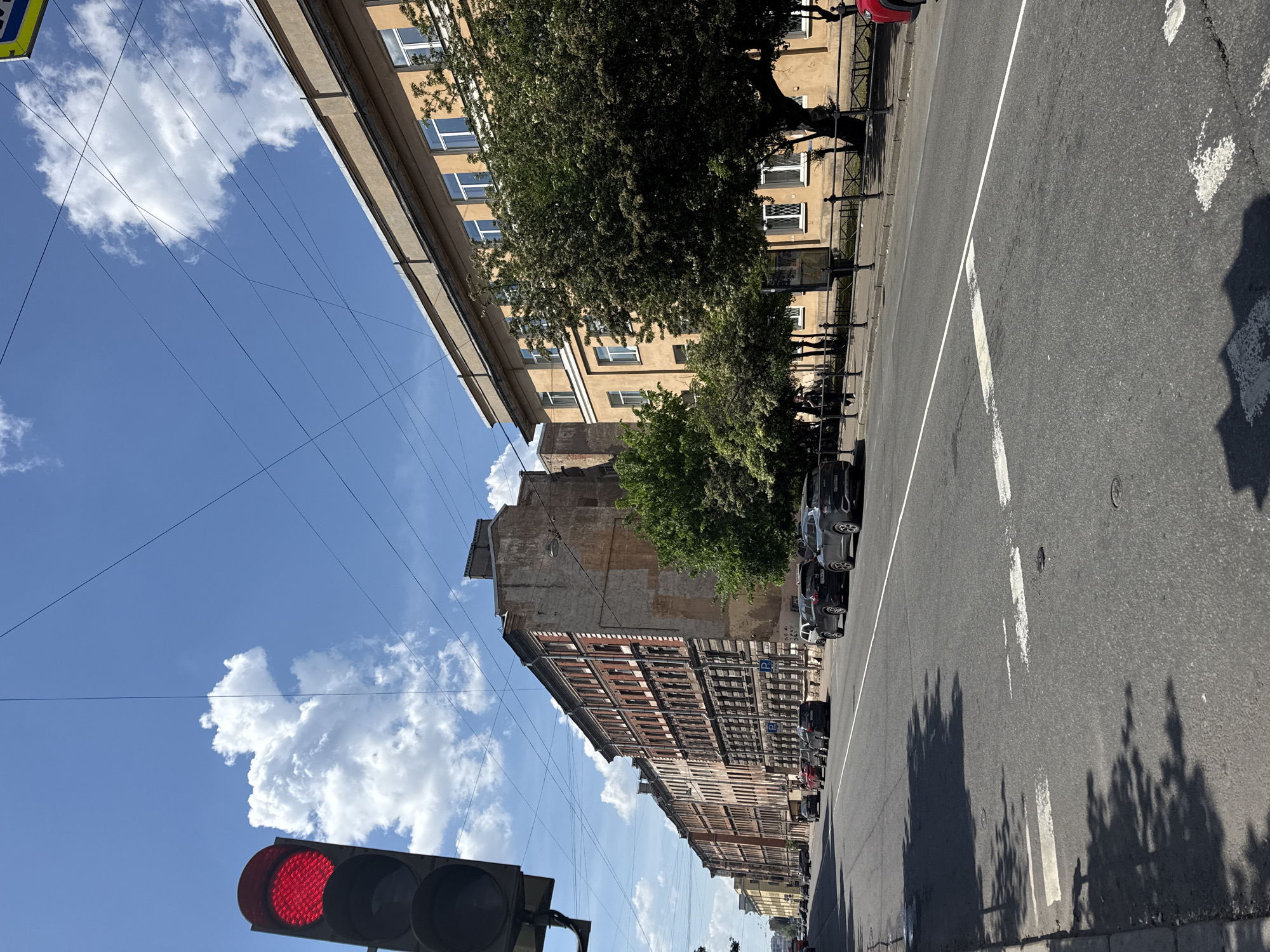 The image displays a sunlit urban street scene, captured from a slightly low angle, looking upwards along the street. The sky, a vibrant blue with scattered white cumulus clouds, is crisscrossed by numerous thin overhead wires, likely for power or public transportation.
On the left side of the street, a multi-story building with a light-colored facade and many windows rises towards the sky. On the right, a row of older, multi-story buildings with varied architectural styles and textures lines the street. Some buildings show signs of age, with exposed brickwork or weathered facades, alongside others with visible windows and balconies. Lush green trees are planted along the right sidewalk, casting long, dark shadows across the asphalt road, suggesting bright sunshine, likely in the mid-morning.
The asphalt road features distinct white lane markings, including a solid white line and dashed lines. Several cars are parked neatly along the right-hand curb, parallel to a black metal fence or railing that separates the sidewalk from the buildings. Small blue signs with a white 'P' symbol, indicating parking, are visible along this railing.
In the lower-left foreground, a traffic light is clearly visible, with its top signal illuminated in red. No people are discernible in the scene. The overall impression is that of a calm, sun-drenched city street on a clear day.