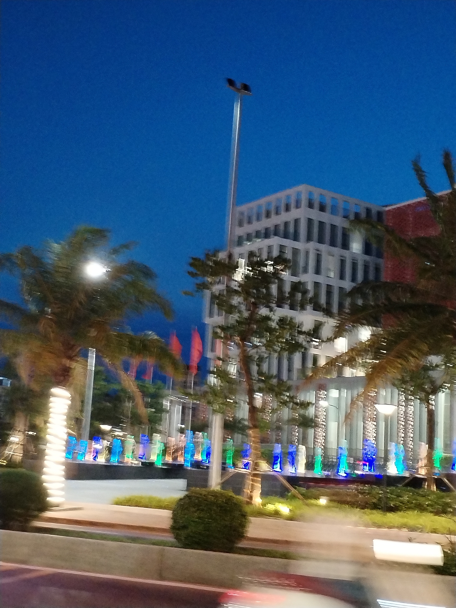 The image shows an outdoor scene in Nha Trang Ward, Vietnam, at dusk or night. The sky is a deep blue. In the foreground, there are palm trees and bushes, and possibly a vehicle moving by. There is a row of illuminated, translucent blue, green, and white figures along a walkway. In the background, there is a tall, modern building with a grid-like facade. A tall streetlight is visible near the building. Red flags are visible in the background.