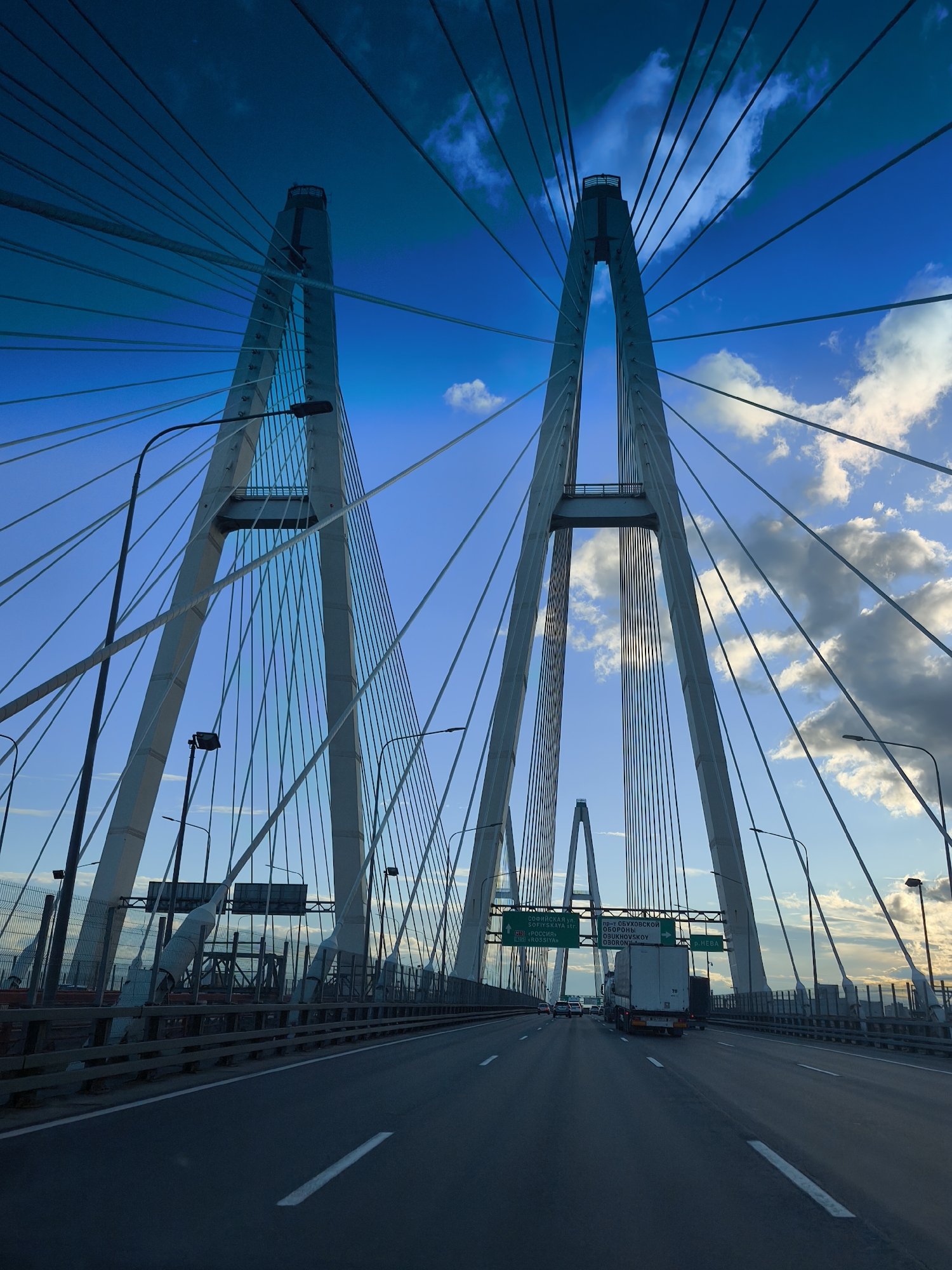 This is a low-angle, wide shot of a cable-stayed bridge against a bright blue sky with scattered clouds. The perspective is from the road, looking forward and upward at the bridge's iconic A-frame towers and numerous suspension cables. The bridge structure dominates the frame, with the road and a few vehicles visible in the foreground and middle ground. Traffic signs are mounted overhead. The lighting suggests either early morning or late afternoon. The overall impression is one of a modern, engineered marvel set against a dramatic sky.