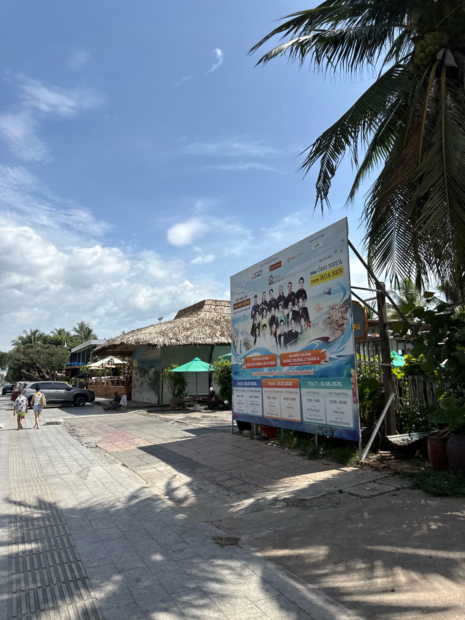 The image depicts a sunny day scene on a sidewalk in Nha Trang, Vietnam. A large billboard, likely advertising an event or promotion, dominates the right side of the frame, featuring images of people and text in Vietnamese. To the left of the billboard, a street with parked cars and pedestrians walking is visible, with buildings featuring thatched roofs in the background, suggesting a coastal or resort area. Palm trees, characteristic of tropical climates, frame the upper right corner of the image. The environment appears to be a public space, possibly a walkway or promenade, with paving stones and shadows cast by the sun.