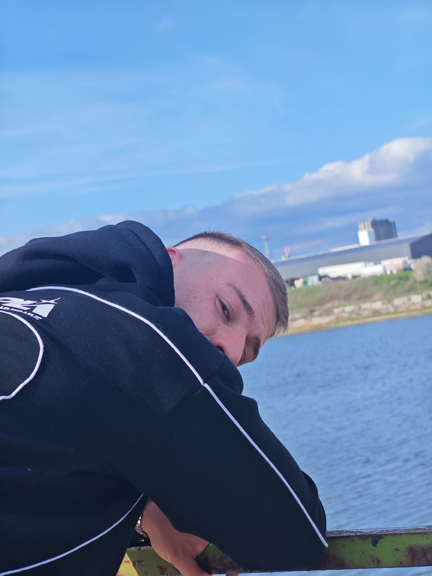 The image shows a fair-skinned young man with a short, faded haircut. He is wearing a black hooded sweatshirt with white piping and is leaning on a green-painted metal railing. Behind him, there is a body of water, likely a lake or river, with a grassy shoreline in the background. Further back, there are industrial buildings and a tower visible in the distance. The sky is blue with scattered clouds.
Given the skyline features, including the tower, and industrial buildings, it is plausible this photograph was taken near the shoreline of a lake or river in the vicinity of Shumen, Bulgaria.