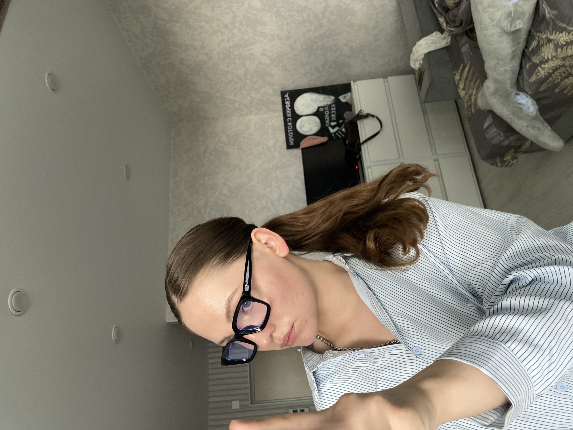 A young woman with brown hair pulled into a ponytail, wearing black-rimmed glasses and a light blue pinstripe button-down shirt, is holding up her left index finger towards the camera. She is looking to her left, away from the camera, with a neutral expression. In the background, a dresser with a television on top is visible, along with a wall decorated with textured wallpaper. A plush toy whale lies on a bed in the lower right corner of the frame. The scene appears to be an indoor residential setting, possibly a bedroom or living area. There is no visible indication of the city of Izhevsk, Russia.