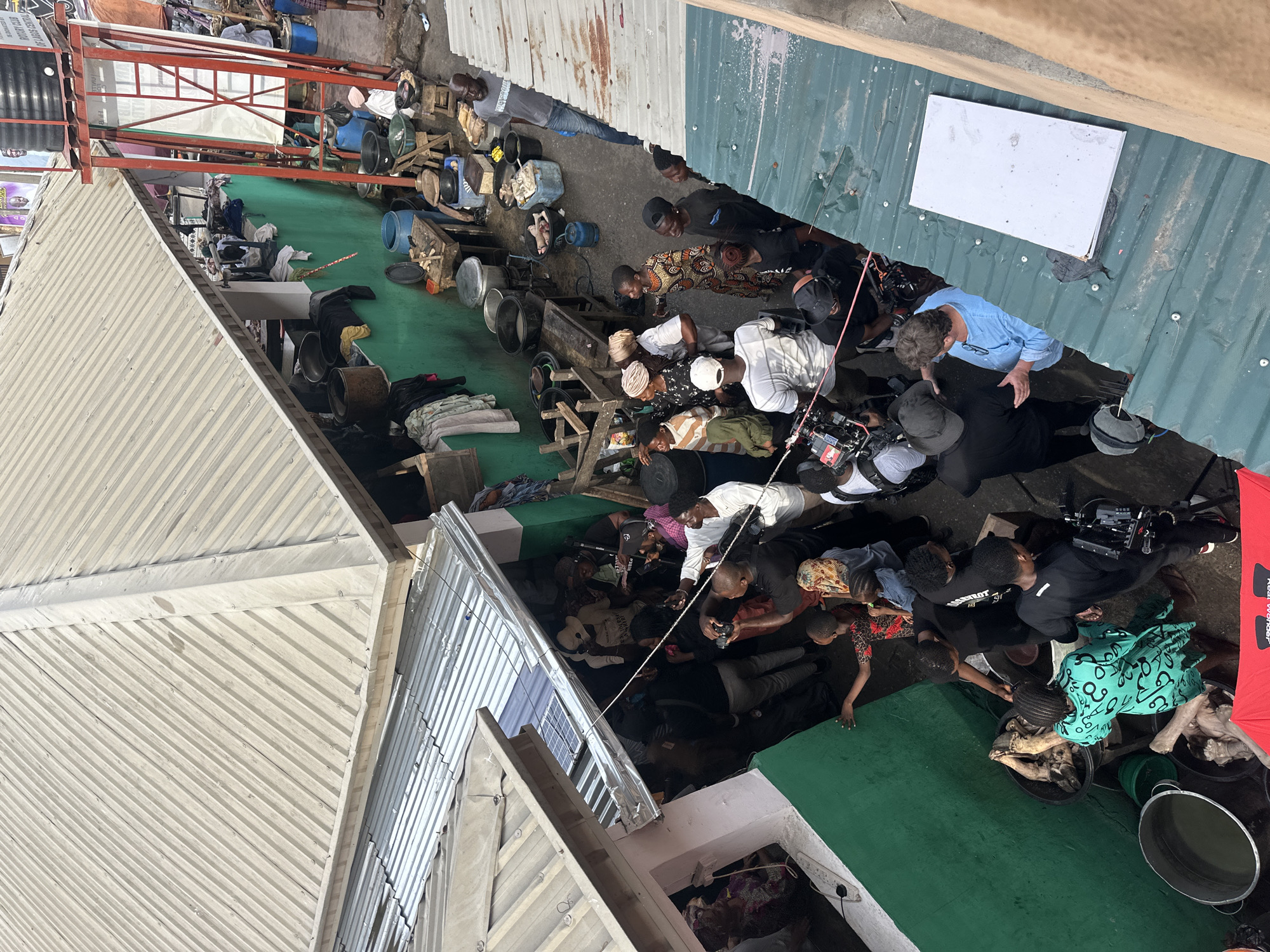 This overhead image captures a busy outdoor scene in an urban area of Lagos, Nigeria, likely an alleyway or a crowded space between buildings. The primary activity appears to be a filming or production event, with numerous individuals gathered around camera equipment.
A large group of people, including men, women, and a few children, populate the scene. Several individuals are operating professional video cameras mounted on rigs, suggesting a film or documentary shoot. One person wears a black t-shirt with "RIVERLAKE" or "RIVERLOT" visible text. Many others are observing the filming, some standing, some crouching.
The environment is characterized by makeshift structures and everyday objects. The ground varies between dark green-painted sections and rough concrete or earth. Corrugated metal roofs, both light grey and dark green, form the overhead structures, and a red metal scaffolding frame is visible in the background. A white rectangular sign is affixed to one of the green roofs.
Scattered throughout the space are various containers, including numerous metal and plastic buckets, basins, and bowls, some of which hold what appear to be animal carcasses (possibly fish or poultry). Wooden crates, stools, and bundles of fabric or clothing are also present. A red umbrella, branded "Real Wonder," is visible near the camera setup, likely for light diffusion or shade.
The scene is brightly lit, indicating daytime. The overall impression is one of a bustling, active urban setting where professional production work is taking place amidst the daily life of the community.