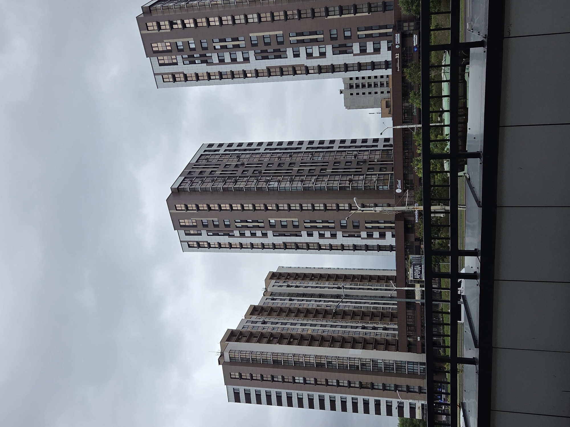 The image depicts an urban landscape dominated by several modern high-rise buildings under a uniformly overcast, light grey sky. The location is Unknown, Unknown.
In the foreground, a black metal railing with vertical bars and horizontal top and bottom rails extends across the lower portion of the frame, suggesting an elevated viewpoint such as a bridge or overpass. Below the railing is a grey, paneled wall or structure.
Beyond the railing, a strip of dark green foliage (trees or bushes) is visible, separating the viewer from the primary structures. Multiple white streetlights on dark poles are interspersed within this area.
The midground and background are dominated by three prominent, multi-story residential towers. These buildings feature contemporary architecture with facades composed of alternating sections of brown/tan and white/light grey panels. Numerous rectangular windows, many with a warm, reflective tint, are visible, some incorporated into enclosed balconies or loggias. The central and rightmost buildings appear to be distinct towers, while the leftmost building is a longer structure. These towers are connected at their base by a two-to-three story commercial podium with darker brown facades.
Several illuminated signs are visible on the commercial levels of these buildings, featuring Cyrillic and Latin text. Notable signs include "АРЕНДА" (FOR RENT) in large white letters, "WB", "КОФЕЙ" (COFFEE), "Dzerzhinskogo 32", "PRADA", "ФАНТОМ" (PHANTOM), and "Маяк" (MAYAK).
In the far background, behind the main high-rises, a plainer, multi-story building with a lighter grey facade, possibly under construction or with minimal detailing, is partially visible. No individuals or vehicles are clearly identifiable, though a dark vehicle is partially obscured by the railing on the right. The overall lighting suggests an overcast or heavily cloudy day.