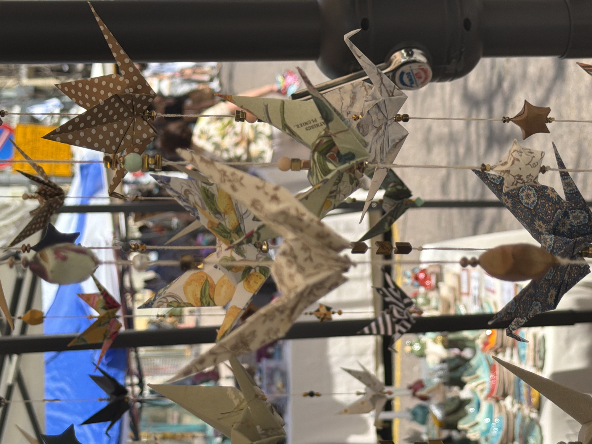 The image captures a vibrant, handmade mobile or garland featuring numerous origami figures, predominantly birds (cranes) and stars, strung on horizontal lines. The display hangs in the foreground, with a shallow depth of field rendering the background significantly out of focus.
The origami creations exhibit a diverse range of patterns and colors. Notable designs include brown paper with white polka dots shaped into a star, light green or beige paper adorned with botanical prints such as lemons and leaves, grey and white patterned paper, intricate dark blue patterned paper, and black and white striped paper. Some smaller, multi-colored and floral-patterned origami pieces are also visible. Various beads and wooden elements, including light green, gold-colored, and dark brown beads, are interspersed along the strings, adding to the decorative nature of the display.
In the blurred background, hints of an outdoor market or fair are discernible, with people and stalls suggesting a lively public setting. The bright, natural lighting indicates it is daytime, likely a clear and sunny day. The photograph is taken from a slightly low angle, looking upwards at the hanging decorations. The overall impression is one of intricate craftsmanship and artistic expression, consistent with items found at a craft market.
Visible text includes the word "PLUMIER." and partially "D'ATHENES" on one of the light green/beige origami cranes, indicating the paper might be repurposed from a printed sheet, possibly an antique botanical or educational illustration. No other distinct text is legible due to the strong blur of the background. Given the location context of Tel-Aviv, Israel, this scene likely depicts a local artisan market.