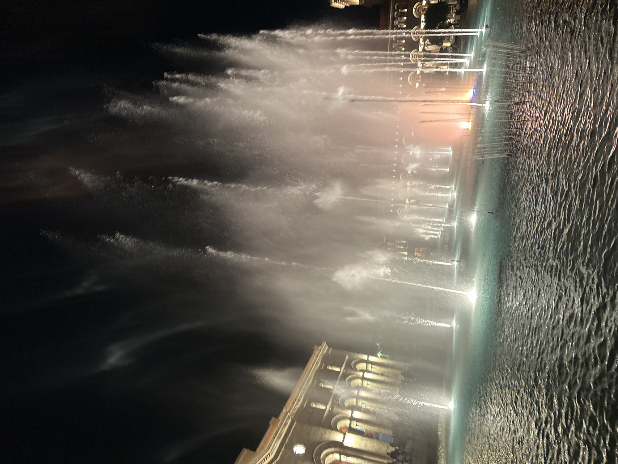 The image depicts a nighttime scene of a large fountain with illuminated water jets. The fountain is located in front of an ornate building with arched windows and lit interiors, suggesting a public square or plaza. In the background, there are additional structures with decorative lighting. The water is moving rapidly, and the air is filled with mist from the fountain. The water's surface appears textured and reflective. The location is Yerevan, Armenia.