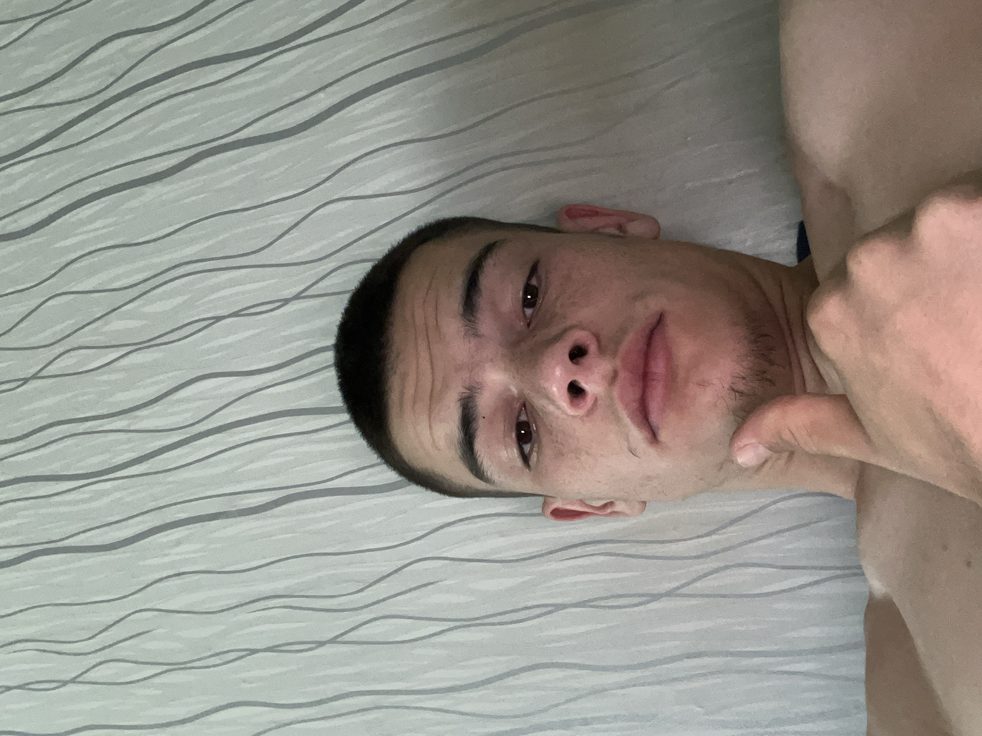 A young man with a short buzz cut and freckles on his face is looking at the camera in a close-up selfie. He is lying down, with his thumb raised in a "thumbs up" gesture. The background is a wall with a grey, wavy pattern. His bare shoulders and chest are visible. The image was taken indoors, in Sopot, Bulgaria.