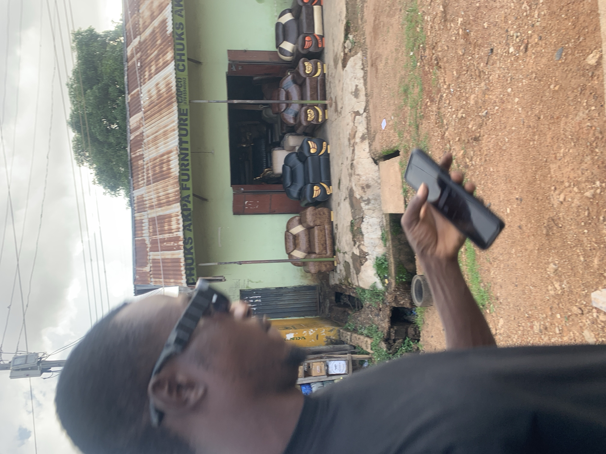 This media file shows a street view in Jos, Nigeria. The scene is dominated by a furniture shop with a corrugated iron roof. The shop's facade is painted light green, and text on the roof reads "AKPA FURNITURE CHUKS AK". Inside the shop, several upholstered chairs are visible, some in dark brown and others in black and cream. In front of the shop, more chairs are displayed, including a prominent dark brown armchair with gold accents.
To the right of the frame, a person is holding a black smartphone in their right hand, with their arm and shoulder visible. The ground is a mix of dirt and some patches of grass, with a small drain visible on the right side. The sky is overcast, suggesting a cloudy day. The overall setting appears to be an outdoor commercial area, possibly a roadside market or street with local businesses. The image is oriented vertically, as if taken with a phone held in portrait mode.