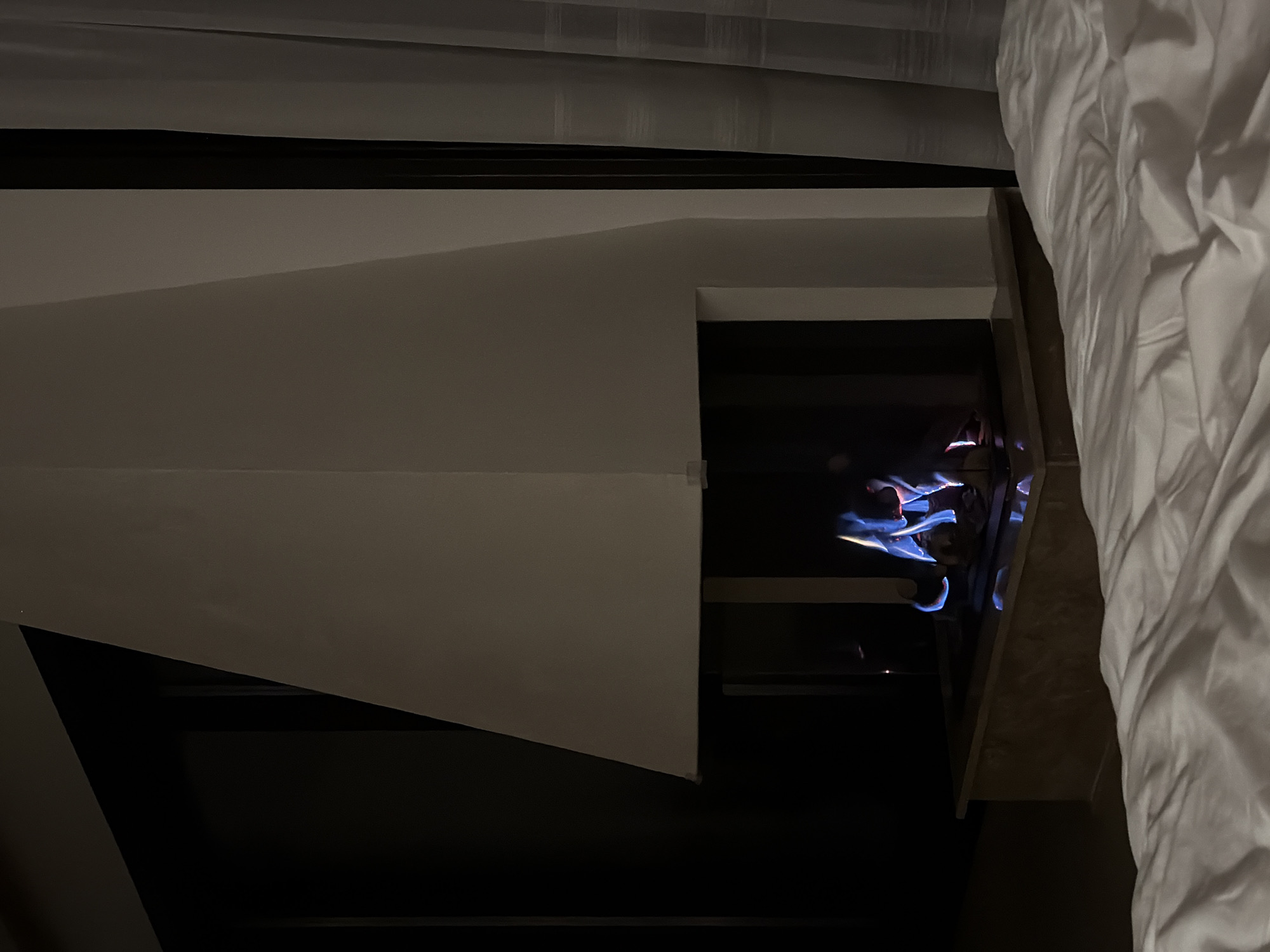 The image depicts an indoor scene with a modern fireplace as the central focus. The fireplace is a substantial, angular structure with a light-colored, possibly white, exterior. Inside the firebox, visible flames flicker with blue and red hues, indicating an active fire. The fireplace is set into a dark, recessed area, with a polished, dark-colored hearth. In front of the fireplace, a bed with white linens is visible in the foreground, suggesting a bedroom or living space. To the right of the fireplace, sheer white curtains are partially drawn, revealing a dark vertical element behind them, possibly a window frame or door. To the left of the fireplace, a dark, solid surface extends upwards, with a diagonal line suggesting the edge of a wall or furniture. The overall lighting is dim, with the fire providing the primary source of illumination. There are no people visible in the image. The location is described as Krasnovo, Bulgaria, implying this is an interior view within that geographical area.