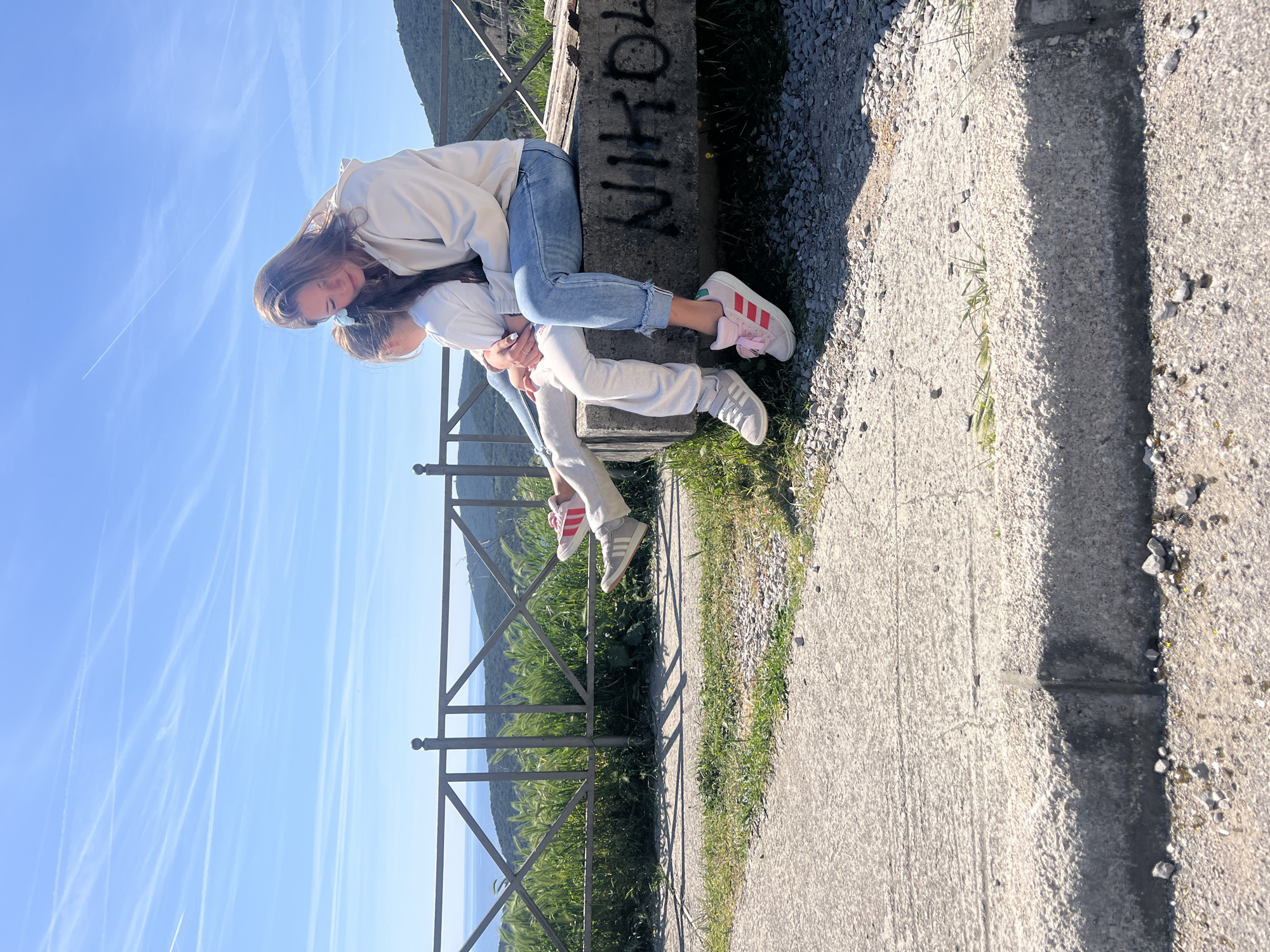 Two individuals, presumably a mother and child, are interacting on a concrete structure. The taller figure, a female with long brown hair, is wearing a light-colored jacket, blue jeans, and pink sneakers. She is leaning over a shorter figure, a child in white attire and grey sneakers. They are positioned on a concrete wall with graffiti ('NIKOLA') visible on its side. A metal railing is nearby, with a view of green foliage and a body of water in the background. The location is in Prijevor, Montenegro. The weather appears sunny and the sky is clear with contrails.