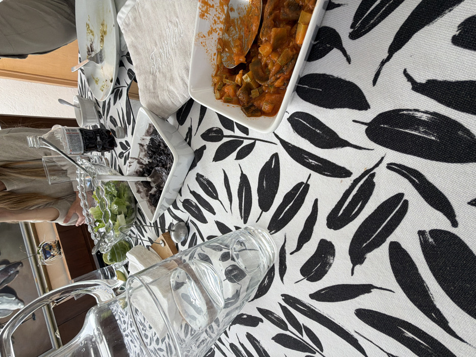 A table with a black and white leaf-patterned tablecloth features a selection of dishes. Square white bowls contain stewed vegetables and a dark, possibly berry-based, dessert. A salad sits in a clear glass bowl. A large clear glass pitcher is in the foreground, angled toward the viewer. In the background, partially visible, are a plate and other serving dishes, along with glimpses of people and a reflection of artwork.