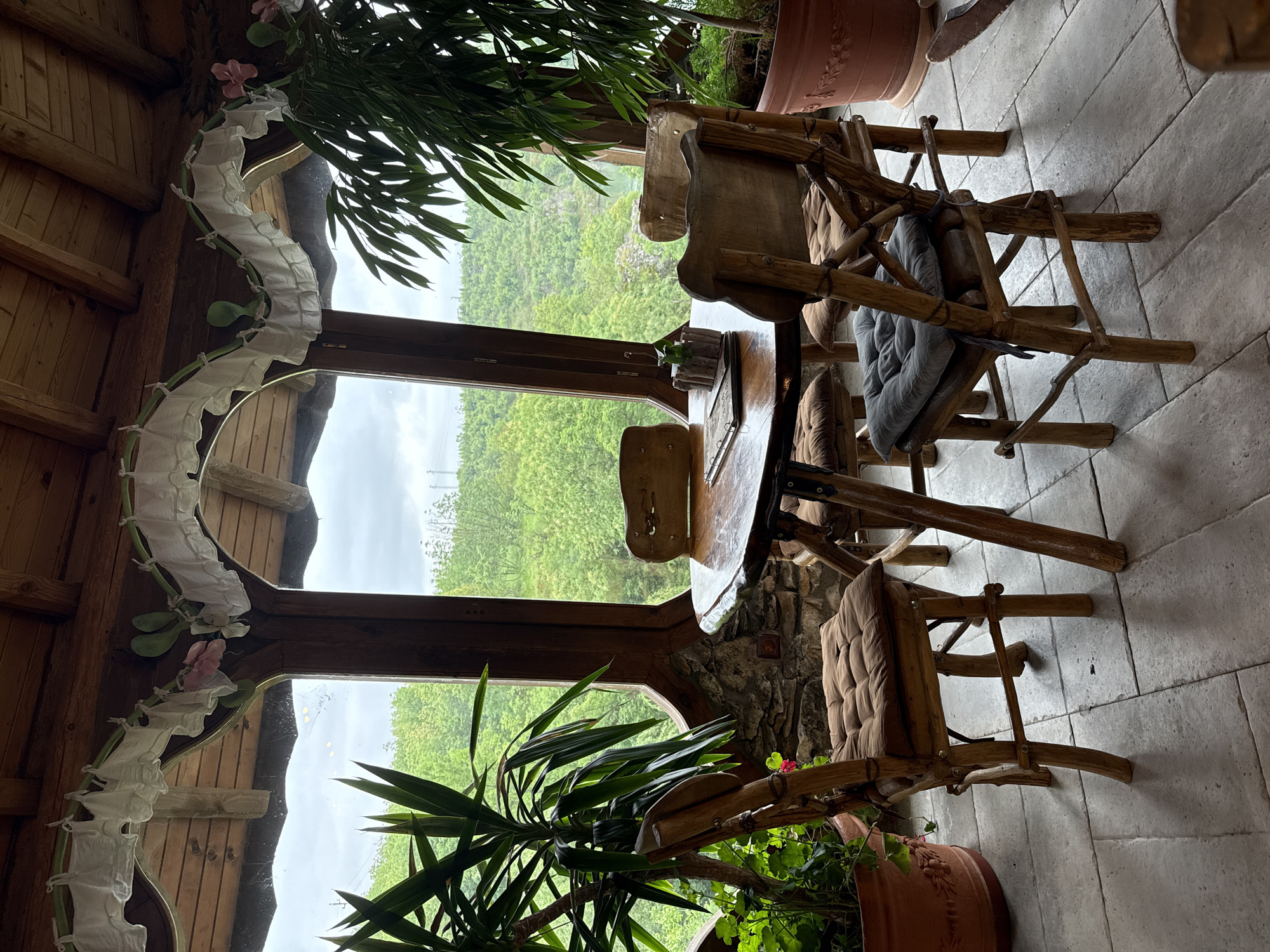The interior of a rustic dining area is shown. A round wooden table is surrounded by chairs made of natural wood branches. Cushions are placed on the chairs. A window provides a view of lush green hills. The ceiling is made of wooden beams and a decorative garland adorns the window frame. Potted plants are present near the window. The floor is tiled with light-colored stone. The scene appears to be located in a nature-oriented setting, possibly near the city of Lukovit, Bulgaria, given the landscape and architectural style.