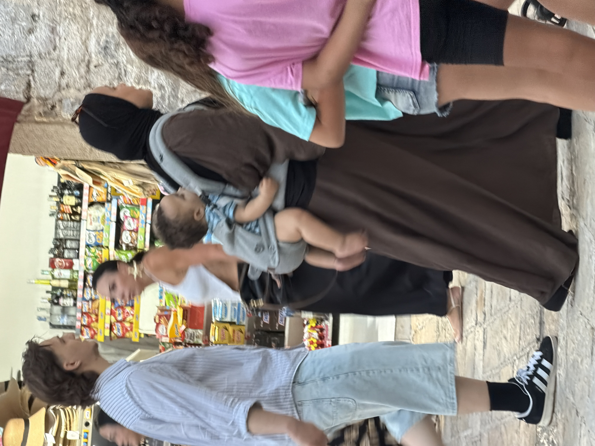 A diverse group of people walk along a cobblestone street in Budva, Montenegro, with a store featuring shelves of merchandise visible in the background. On the left, a man with light brown hair and a light blue striped shirt walks forward, his hands in his pockets. He wears light blue, loose-fitting shorts and black sneakers with white laces and three white stripes. To his right, a woman wearing a white tank top and large gold earrings walks away from the camera, her dark hair pulled back. She wears dark pants. In the center, a person wearing a dark brown abaya and a black hijab carries a baby in a carrier on their front. The baby wears a light blue and grey outfit. To the right of the abaya-clad person, a young girl with curly brown hair wears a bright pink t-shirt, and a second young girl with curly brown hair wears a turquoise t-shirt and denim shorts. The store behind the people has rows of packaged goods, including snacks and drinks, displayed on shelves.