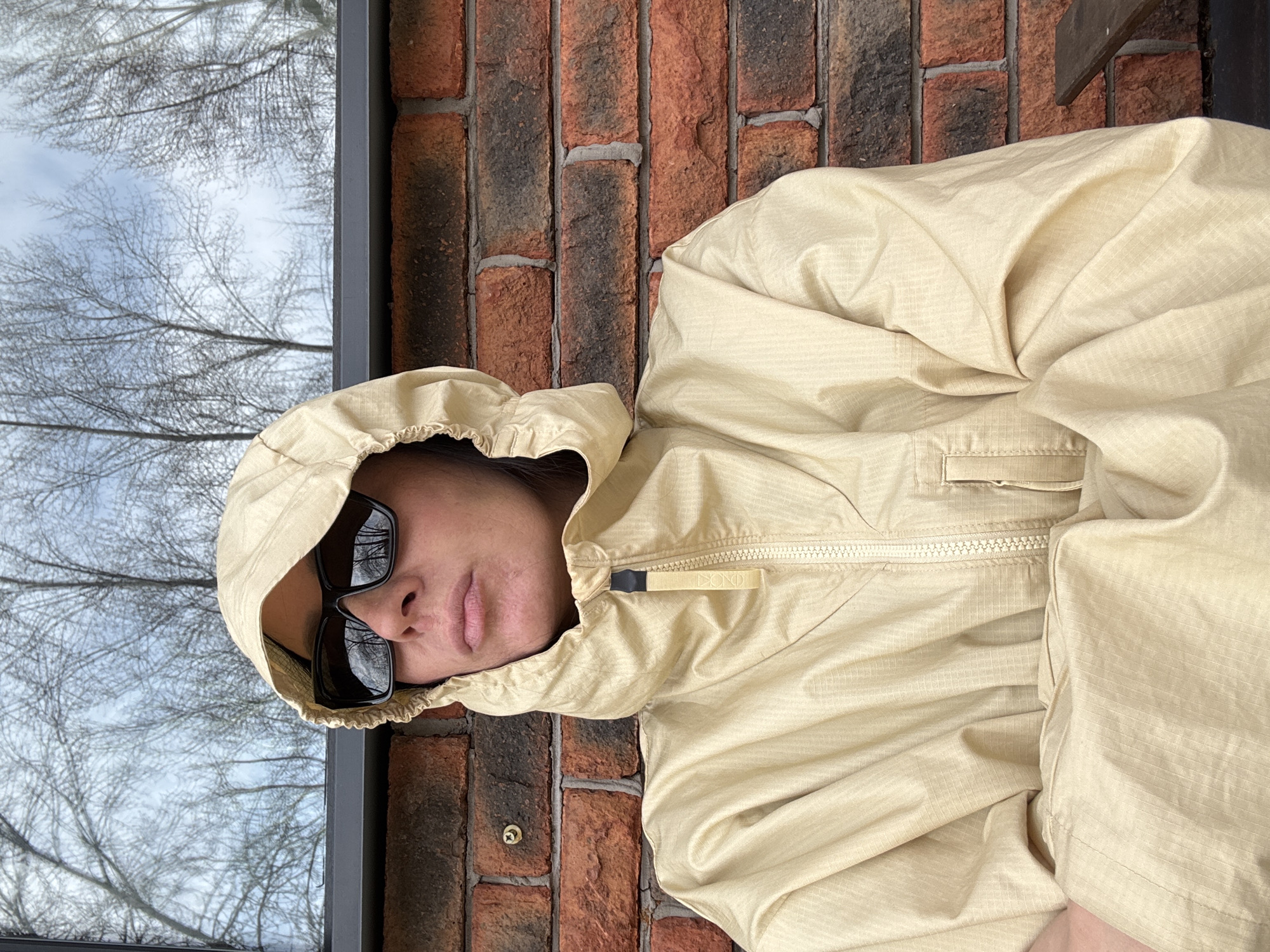 A person wearing a light beige hooded jacket and dark sunglasses is lying near a brick wall. A window is visible to the left, reflecting tree branches and a cloudy sky. The setting is Chaikovsky, Russia, indicated by the wall and window location.