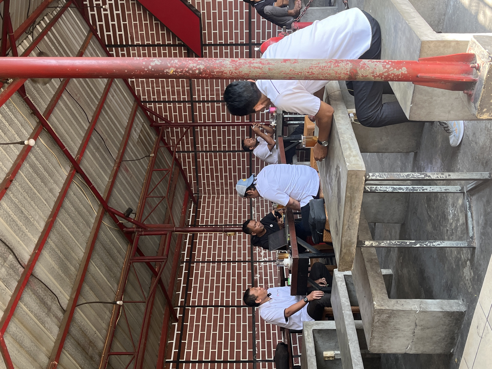 In a covered outdoor seating area in Singosari, Indonesia, five men are gathered at concrete tables and benches. The seating area is open on the sides, with a corrugated metal roof supported by red metal beams and poles. The back wall is constructed of red brick with white mortar lines. In the foreground, a man in a white polo shirt and dark pants sits with his back to the camera, facing a concrete table. To his left, another man in a white polo shirt and dark pants is seated at the same table, looking at his phone. Further left, two men are seated at a wooden table. One, wearing a white polo shirt, is speaking, while the other, in a black t-shirt, is looking at a laptop. To the right, a man in a white t-shirt and a blue cap is seated at a table, facing away from the camera. To the far right, a person's legs are visible sitting on a bench. On the tables, there are glasses of iced tea, cigarettes, and a laptop. The overall atmosphere appears casual, with patrons engaged in conversation and activities.