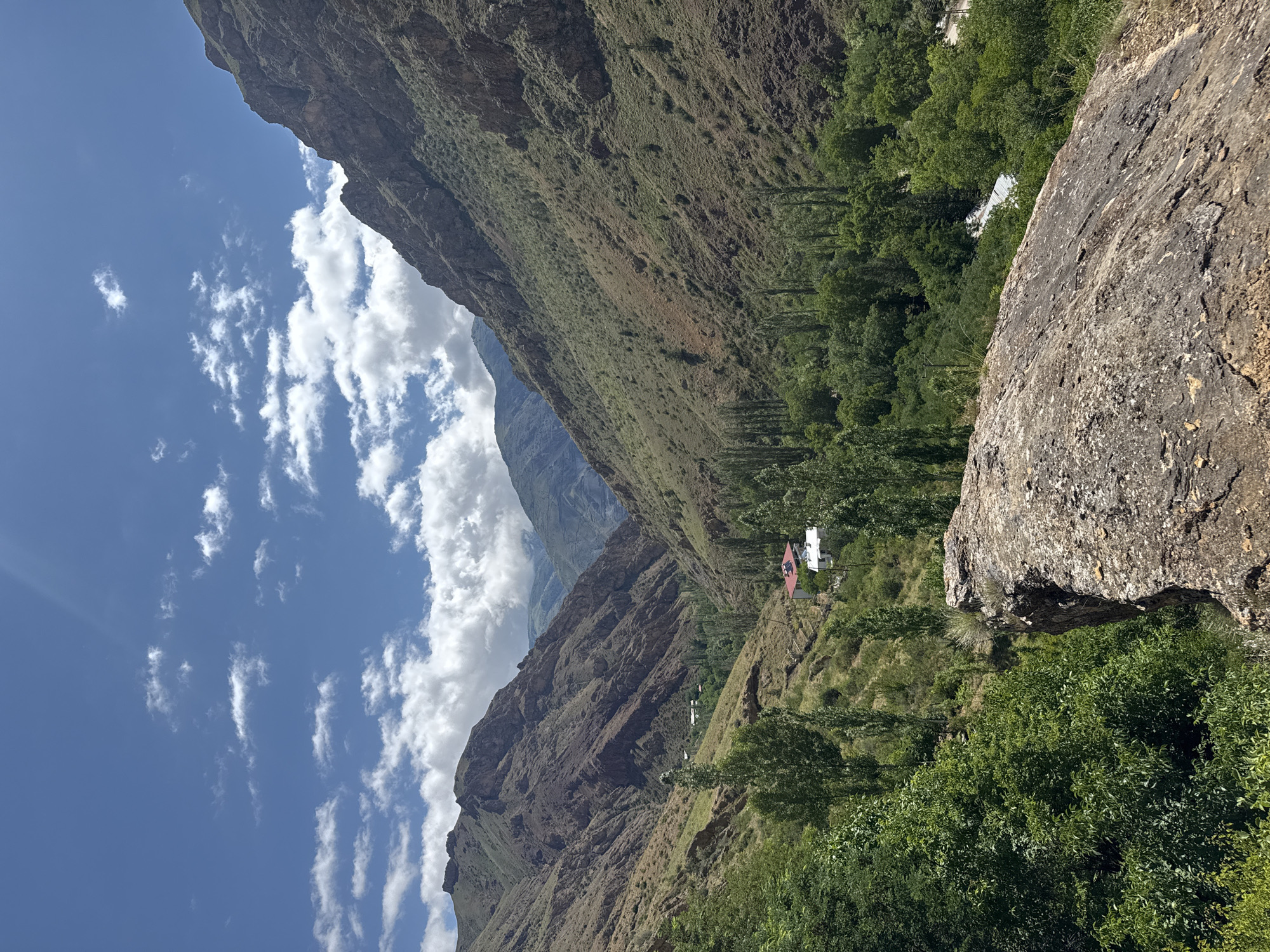 The image depicts a scenic view of a valley in Tortum, Turkey. The foreground features a large, textured rock in the lower right quadrant. Beyond the rock, the valley opens up, revealing steep, brown, and green mountainous slopes on either side. A dense cluster of green trees fills the valley floor, interspersed with tall, slender trees that reach towards the sky. Two white buildings with red roofs are visible nestled amongst the trees, suggesting a small settlement. The sky above is a vibrant blue, dotted with fluffy white clouds, and a bright sunbeam casts a glow across the upper left portion of the frame. The overall environment is natural and mountainous, with signs of human habitation in the form of buildings and what appears to be a path or road winding through the trees. The scene is captured on a clear, sunny day.