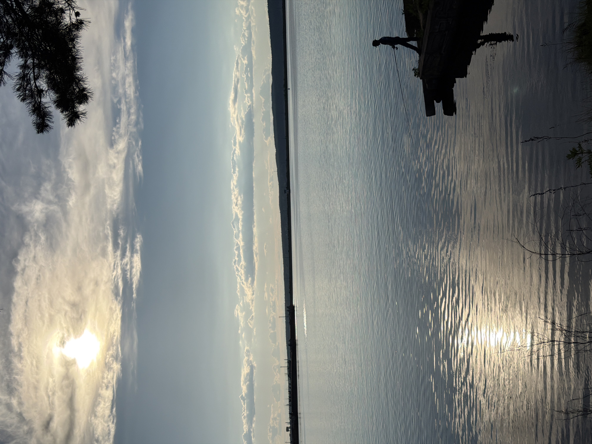 This image depicts a serene outdoor scene, likely captured during sunrise or sunset over a wide body of water, characteristic of the Kama River near Chaikovsky, Russia. The sky, partially cloudy, fills the upper half of the frame. The sun, a bright, diffused orb, is visible in the upper left behind thin clouds, casting a warm glow and illuminating the wispy cloud formations around it.
The lower half of the image features the tranquil water surface, covered in gentle ripples. The sun's reflection creates a shimmering path of light that extends across the water towards the foreground, enhancing the impression of morning or evening light.
On the right side of the foreground, a dark silhouette of a person is visible, standing on what appears to be a small dock or a large log extending into the water. The person is holding a long, thin object, suggesting they are fishing. Dark, silhouetted tree branches, possibly pine, frame the extreme edges of the image on the left and right, indicating vegetation along the banks.
Across the expansive water, a distant shoreline appears as a low, dark, unbroken line, likely forested hills, forming the horizon. The overall lighting and composition evoke a sense of peace and natural beauty. There is no visible text in the image.
