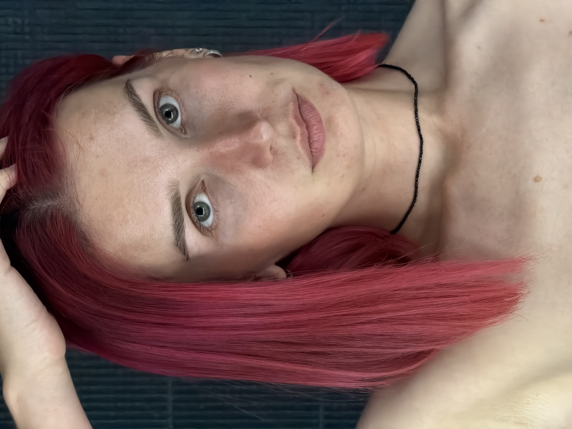 A close-up, waist-up portrait of a young Caucasian female with bright pink shoulder-length hair is shown against a dark, textured background. She is looking directly at the camera with a neutral expression. Her right hand is raised and resting on her head, slightly obscuring the top of her hair. She has light skin with some freckles and blemishes. Her eyes are light green. She is wearing a thin black beaded choker necklace. The image provides no information or visual cues that can be used to determine the location in the city of Chaikovsky, Russia.