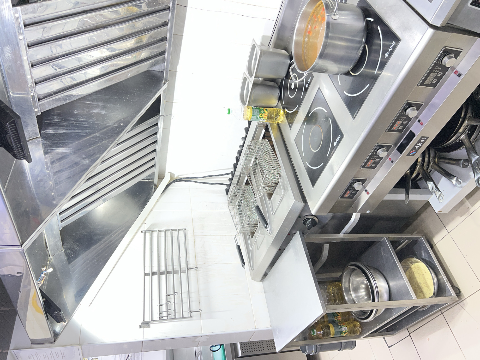 The image depicts a commercial kitchen environment in Plast Univska, Russia. The scene focuses on stainless steel cooking equipment, including a large exhaust hood overhead, a commercial deep fryer with two baskets, and an induction cooktop with a pot of soup on one of the burners. To the left of the cooking equipment is a metal shelving unit holding bottles of cooking oil and several metal bowls, one containing a pale yellow substance. The walls are tiled with white square tiles, and the floor appears to be tiled in a neutral color. No individuals are visible in the image. The lighting suggests an indoor setting, likely illuminated by overhead kitchen lights.