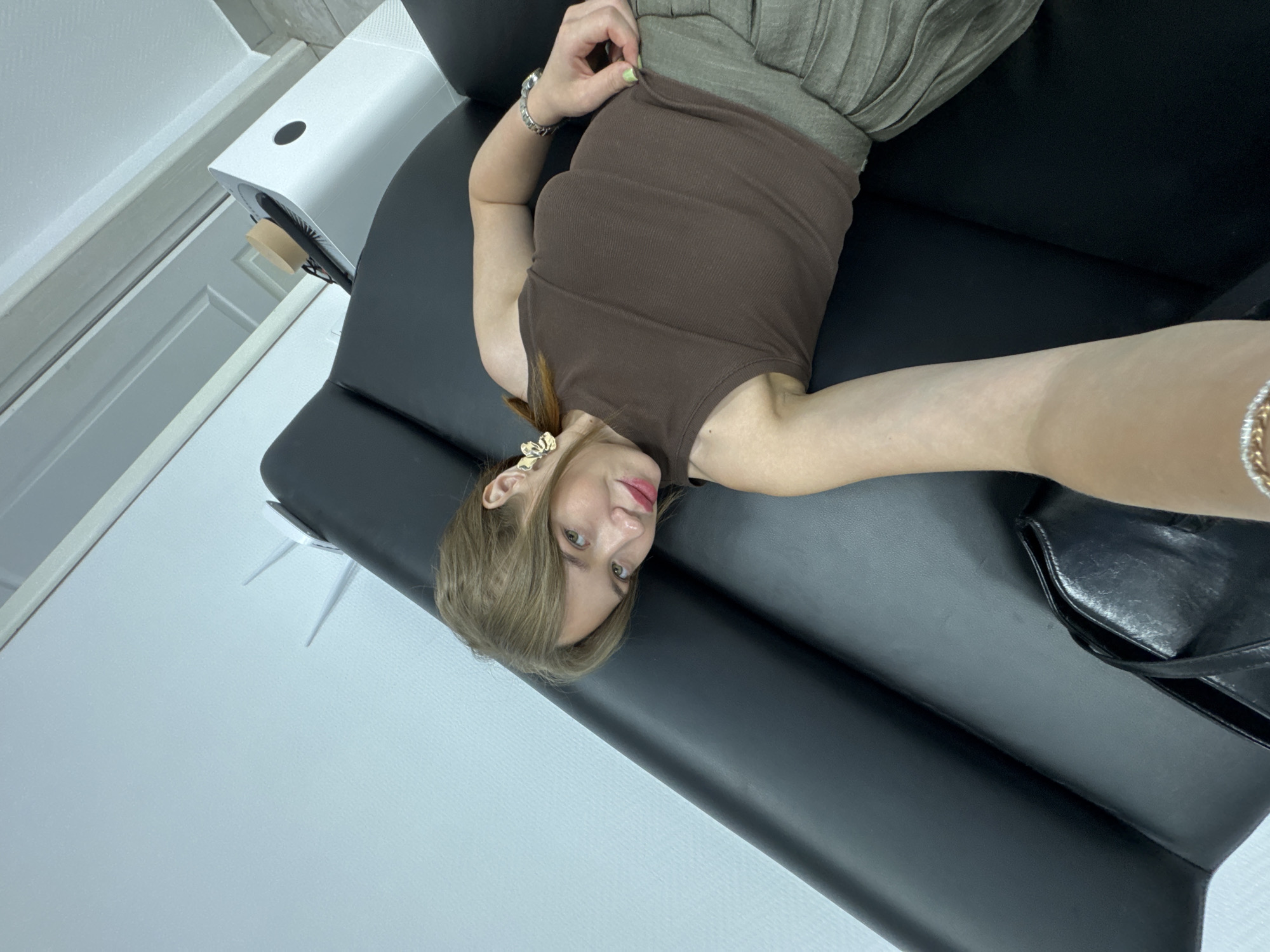 A young woman with light brown hair and fair skin is lying on a black leather couch. She is wearing a brown sleeveless top and a green pleated skirt. She has on large gold earrings and a bracelet on her left wrist. Her right hand is on her skirt. She is holding a phone in her left hand, taking a selfie. A black handbag is to her left on the couch. To the right of the couch, a white air purifier is visible. The background appears to be an indoor setting with white walls and a light blue ceiling. There is also a door frame visible. There are no indicators to suggest this location is in Chaikovsky, Russia.