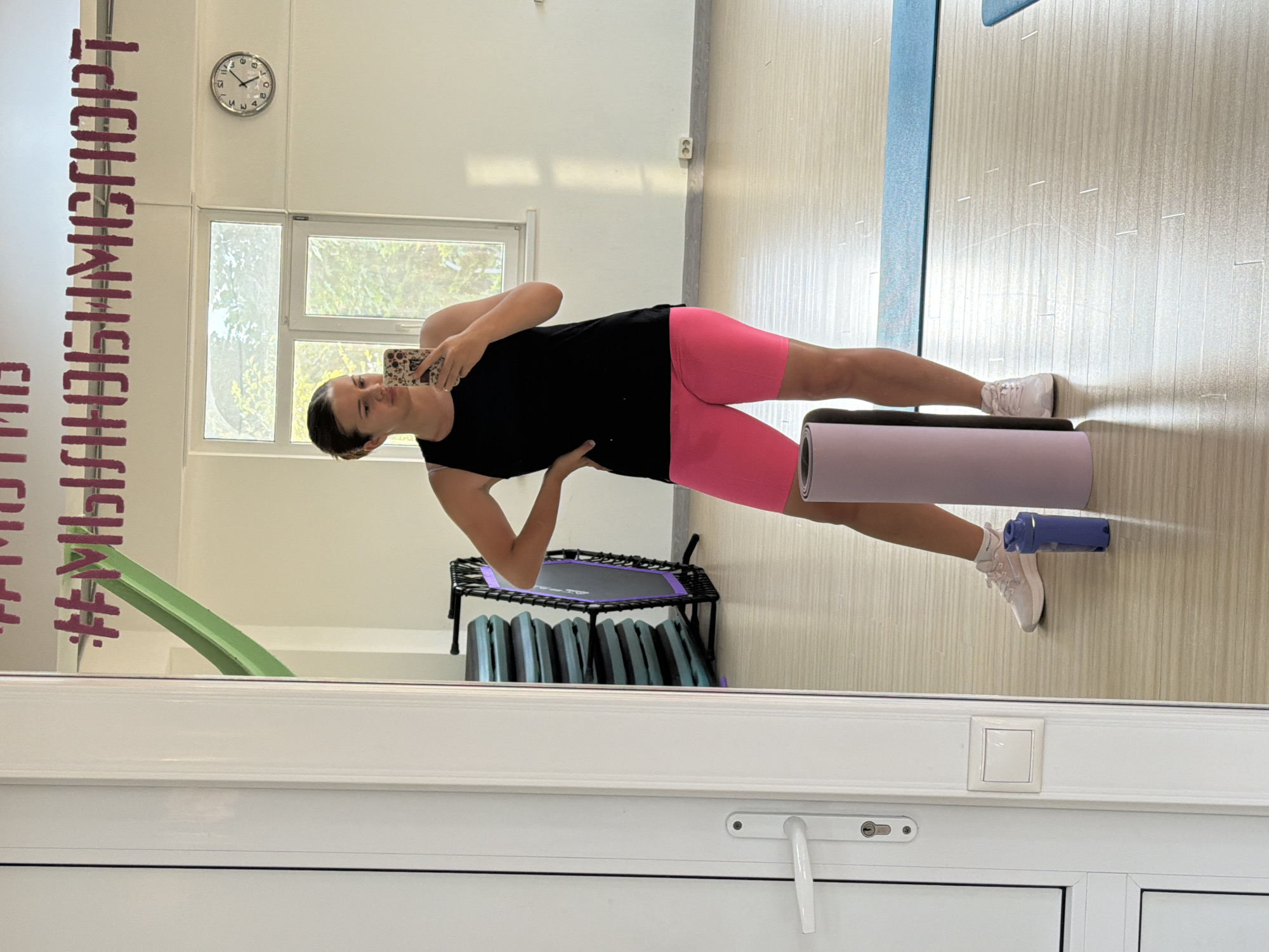 A young woman is captured in a mirror selfie, performing a side plank exercise in what appears to be a gym or fitness studio. She is wearing a black sleeveless top and bright pink bike shorts. Her body is positioned horizontally, supported by her forearm and the side of her foot, with her hips lifted off the floor.
In the background, reflected in the mirror, are gym equipment like a small trampoline and stacked mats. A clock on the wall shows it is approximately 11:07. Light streams in from windows, illuminating the space. A yoga mat is rolled out on the floor next to her, and a blue water bottle is propped against the wall. The overall mood seems focused and determined, as she is engaged in her workout.
Visible text on the mirror, in large, bold, dark pink letters, appears to be a hashtag followed by several words that are difficult to fully decipher due to the reflection and partial visibility, but some letters suggest words like "Finish" and "Champ". The setting is indoors, with white walls and a light-colored floor. The scene suggests a daytime workout session.