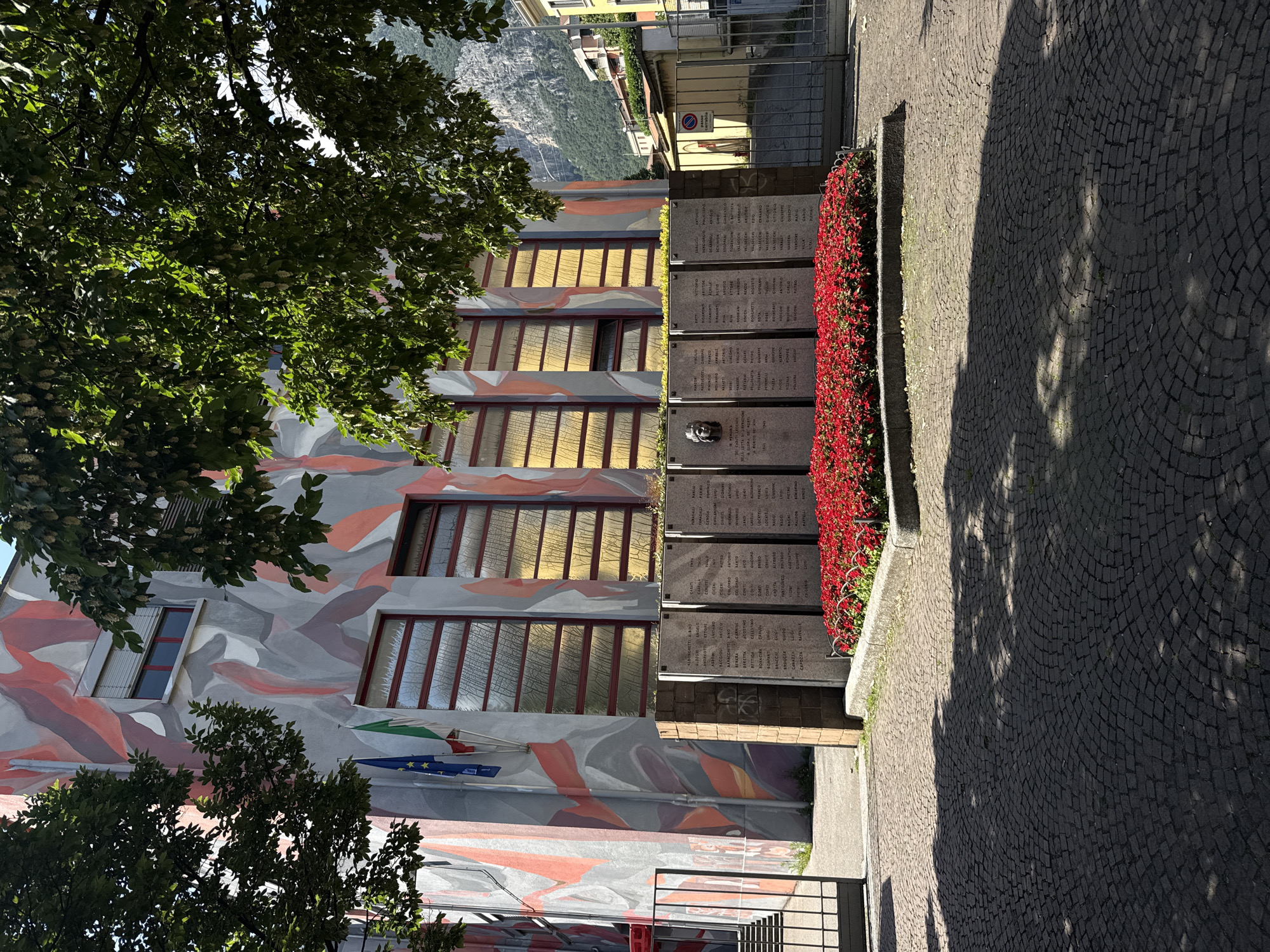 The image depicts a war memorial in front of a building with a vibrant, abstract mural. The memorial consists of several stone plaques inscribed with names, arranged in a row and fronted by a bed of bright red flowers. A bas-relief sculpture of a face is visible on one of the plaques. The building's facade features large, multi-paned windows with a reddish frame and blinds. Two flags, one blue with stars and another green, white, and red, are affixed to the building to the left of the memorial. The foreground is paved with cobblestones, casting dappled shadows from overhead trees. In the background, a steep, tree-covered mountainside is visible, along with residential buildings and a metal gate. The scene is brightly lit by sunlight, suggesting daytime. The overall impression is one of solemn remembrance combined with a striking architectural aesthetic.