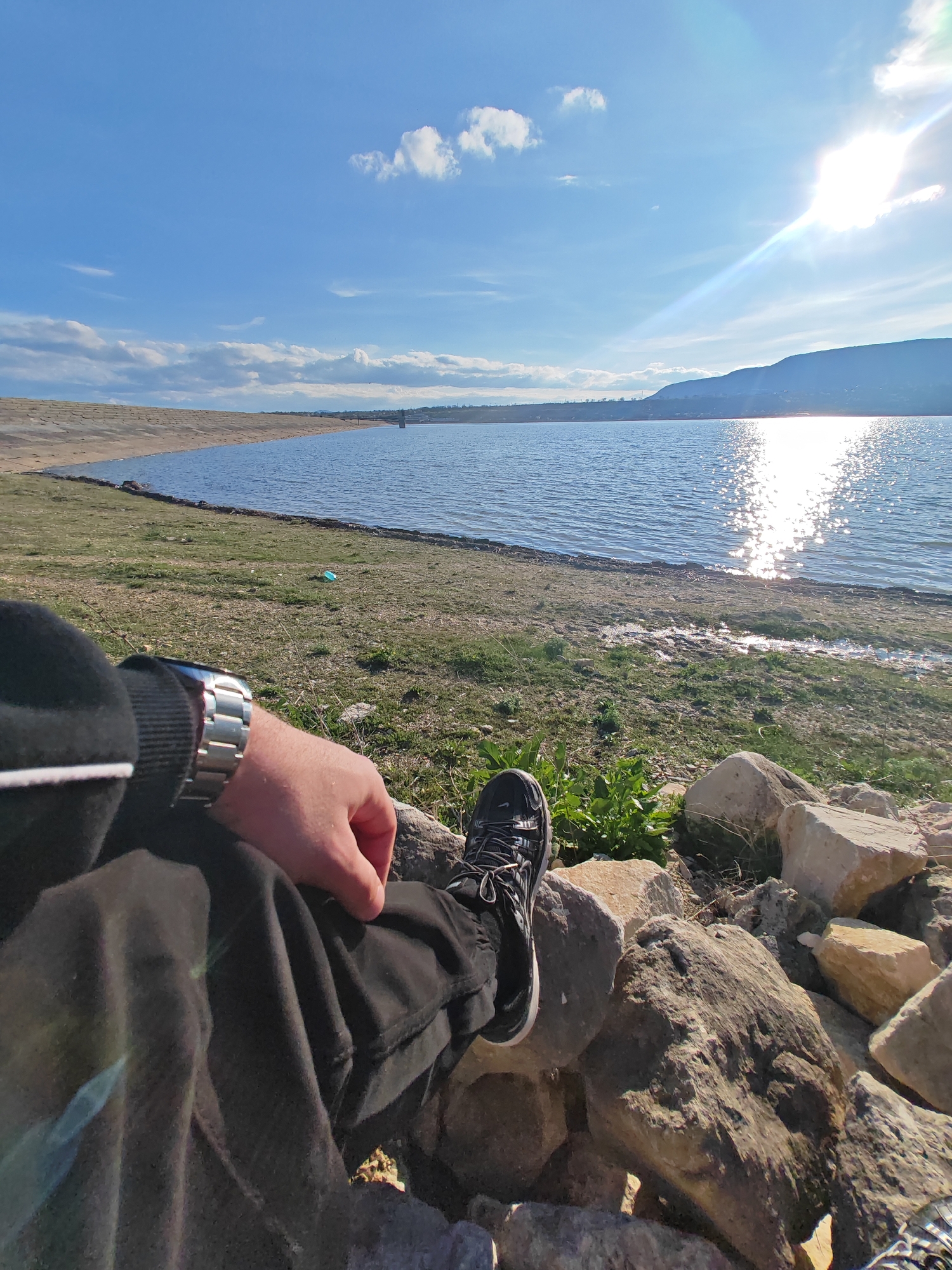 A first-person view image shows a person sitting near the shore of a lake on a sunny day. The person is wearing dark pants and a dark long-sleeved shirt. A silver watch is visible on the person's left wrist. Their right leg, with a black and grey athletic shoe, rests on some rocks. The lake is calm and reflects the sunlight, creating a shimmering effect. In the background, there is a landmass with a hill in the distance and a sandy shoreline. The sky is blue with scattered clouds and a bright sun. The area appears to be a natural landscape. It is unclear whether this location is near the city of Shumen, Bulgaria or is near a different location.