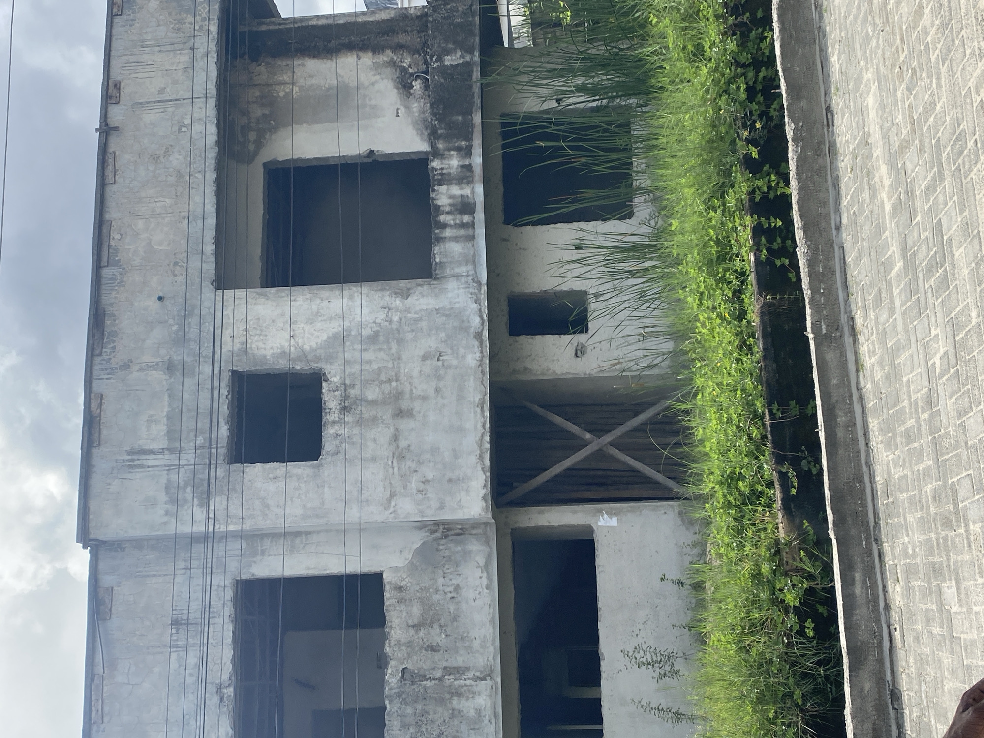 The image depicts an unfinished concrete building with multiple empty window and door openings. The building shows signs of weathering and possible decay. Adjacent to the building is a narrow strip of land overgrown with vegetation. The location is specified as Iranla, Nigeria. No people or actions are visible in the image.