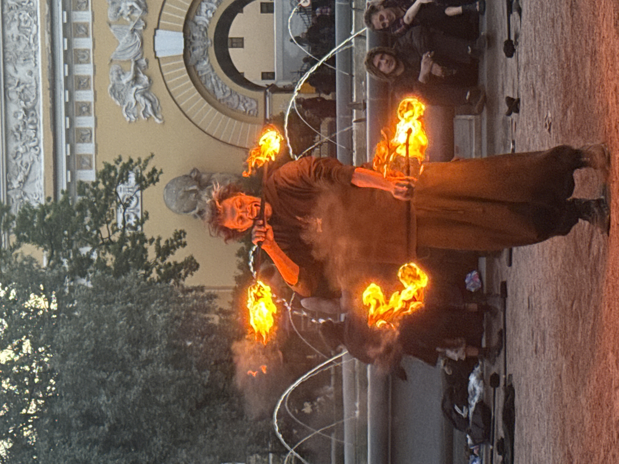 The image depicts a male fire performer in the process of manipulating flames from two torches, one held near his mouth and the other in his hand. He wears a dark, loose-fitting outfit. The background features a fountain with multiple water jets and a building with classical architectural elements, including ornate carvings and an arched entrance. Several spectators are visible in the middle ground, seated and observing the performance. The overall scene suggests an outdoor public space, likely a square or park, within Saint Petersburg, Russia, possibly near an area with fountains and historic architecture, such as Palace Square or the Mikhailovsky Garden.