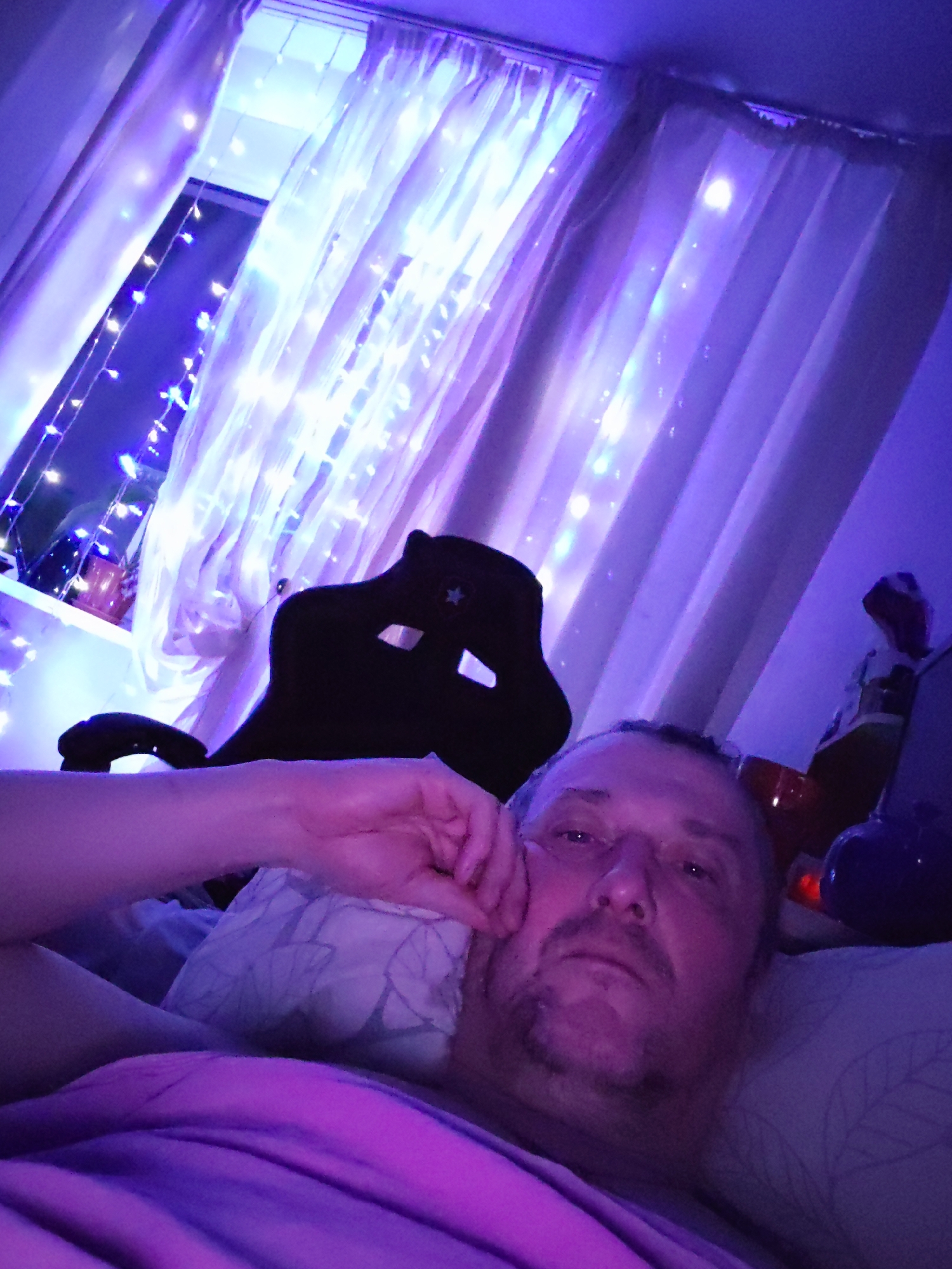 A low-angle indoor selfie shows a middle-aged man lying on a bed. The room is lit with purple and blue lights, including string lights hanging in front of a window, which is behind the headboard of the bed and to the left of the man. The window is draped with sheer curtains. A black gaming chair is positioned behind the head of the bed. A pillow with a leaf-like pattern is behind the man’s head. The man is resting on his left side, and his hand is touching his face. He is wearing a pink shirt. To the right of the bed, there is a shelf with various objects, including a figurine and a blue container. The location is the city of Chaikovsky, Russia.