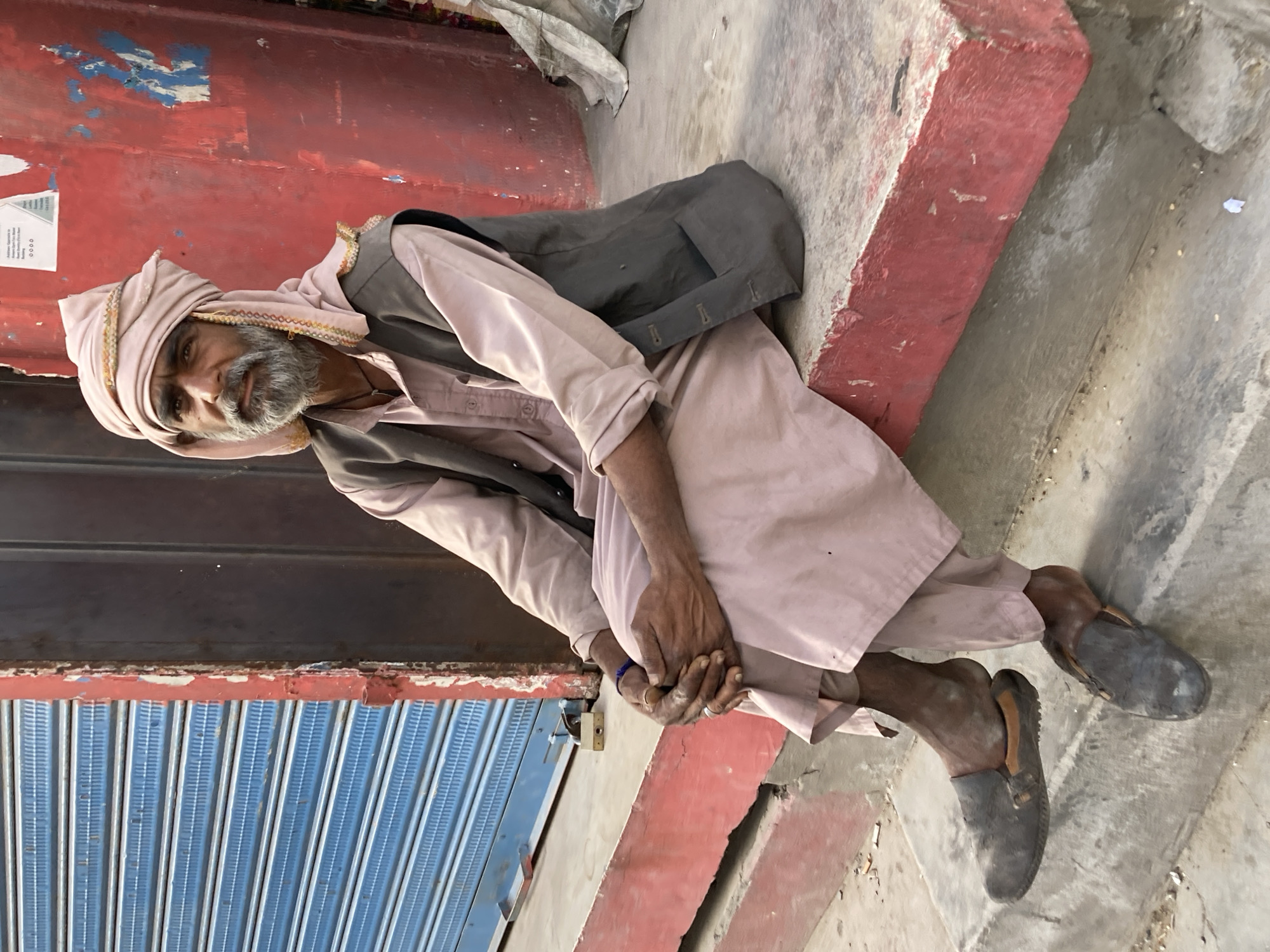 In Quetta City Tehsil, Pakistan, an elderly man wearing a light-colored tunic, a dark vest, a head covering, and slip-on shoes sits on a step. He has a gray beard and holds his hands clasped in front of him. Behind him is a red wall and a blue corrugated metal door. The setting appears to be outdoors during daylight.