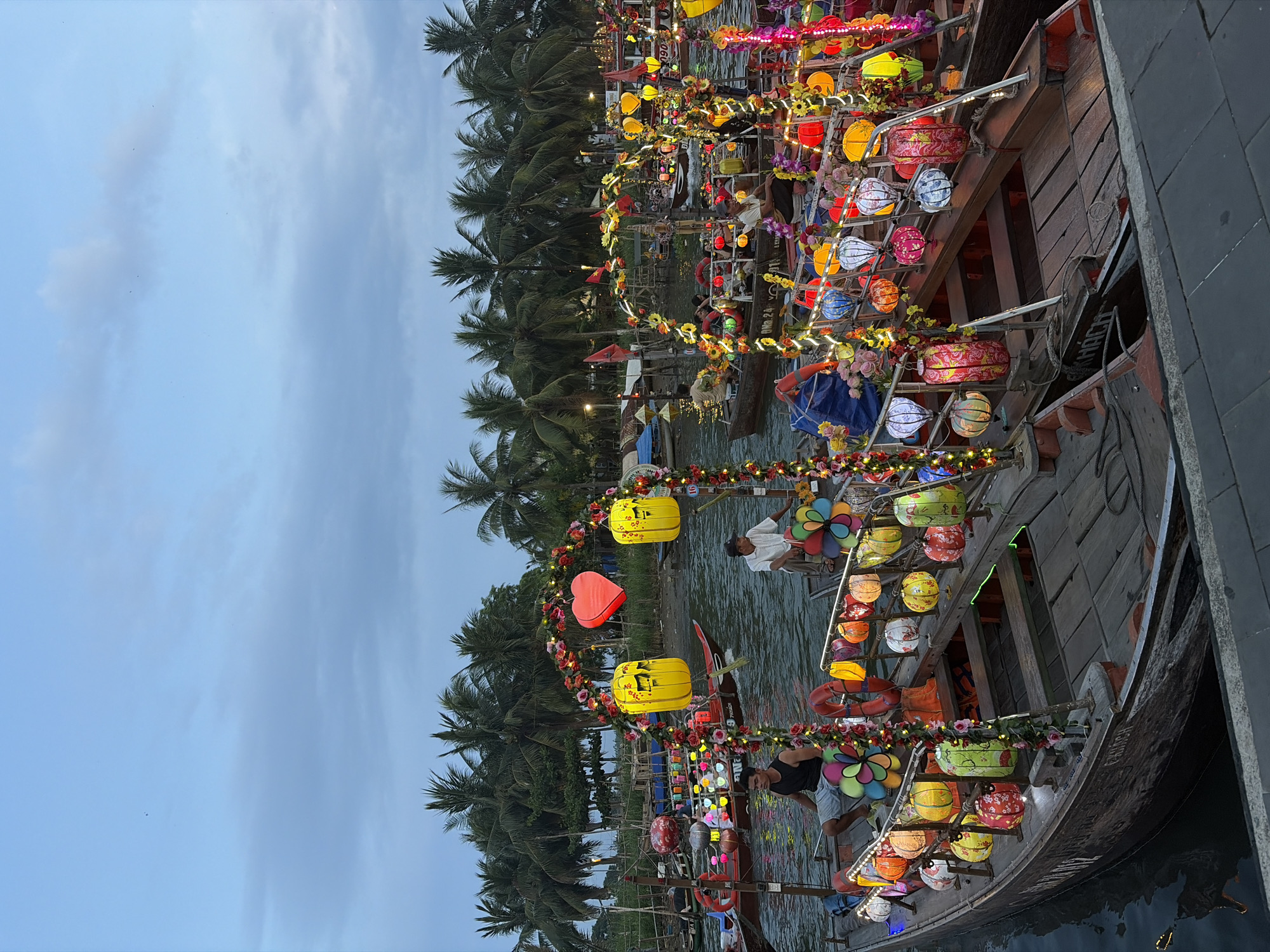 The image depicts a vibrant evening scene on a river or canal in Hội An, Vietnam, showcasing numerous traditional wooden boats elaborately decorated with colorful lanterns and garlands. The sky is a pale blue with soft, diffuse clouds, indicating dusk or early evening, a time when the illuminated decorations become prominent.
The main subjects are the boats, which are adorned with a profusion of spherical, cylindrical, and accordion-shaped lanterns in hues of yellow, red, orange, blue, green, pink, and white. Many lanterns display intricate patterns. String lights and artificial floral garlands, predominantly in reds, pinks, and yellows, drape across the boats' railings and structures. Notable decorations include large yellow lanterns with dark patterns, a prominent red heart-shaped ornament, and multi-colored pinwheel-like elements. Orange life preservers are visible on the sides of some vessels.
Several people are present on the boats. One individual in a white shirt and dark trousers is visible near the front of a boat, and another in a dark shirt is further along, suggesting they are operating or preparing the boats for passengers. The activity appears to be related to evening boat tours or a festive event, as indicated by the extensive decorations.
In the background, a dense line of tall palm trees frames the riverbank, their dark silhouettes contrasting with the lighter sky. The water is calm, reflecting some of the overhead light.
Visible text on one boat includes "OMD 24" and "7900". Another boat displays part of the word "KHÁCH," which translates to "guest" in Vietnamese, reinforcing the interpretation of these as passenger boats. The overall atmosphere is festive and lively, typical of tourist attractions in Hội An at twilight.