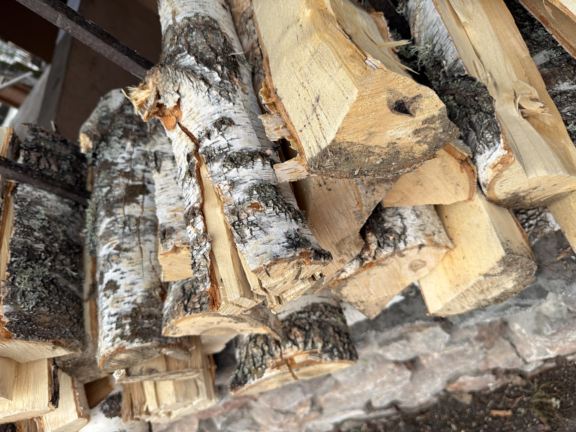 The image shows a stack of chopped firewood, primarily birch, located in Taskino, Russia. The wood is stacked outdoors, seemingly under a partially visible wooden structure. The cut ends of the birch logs show the distinctive white bark and exposed wood grain. The area beneath the firewood appears to be a combination of stone or concrete fragments and exposed soil.
