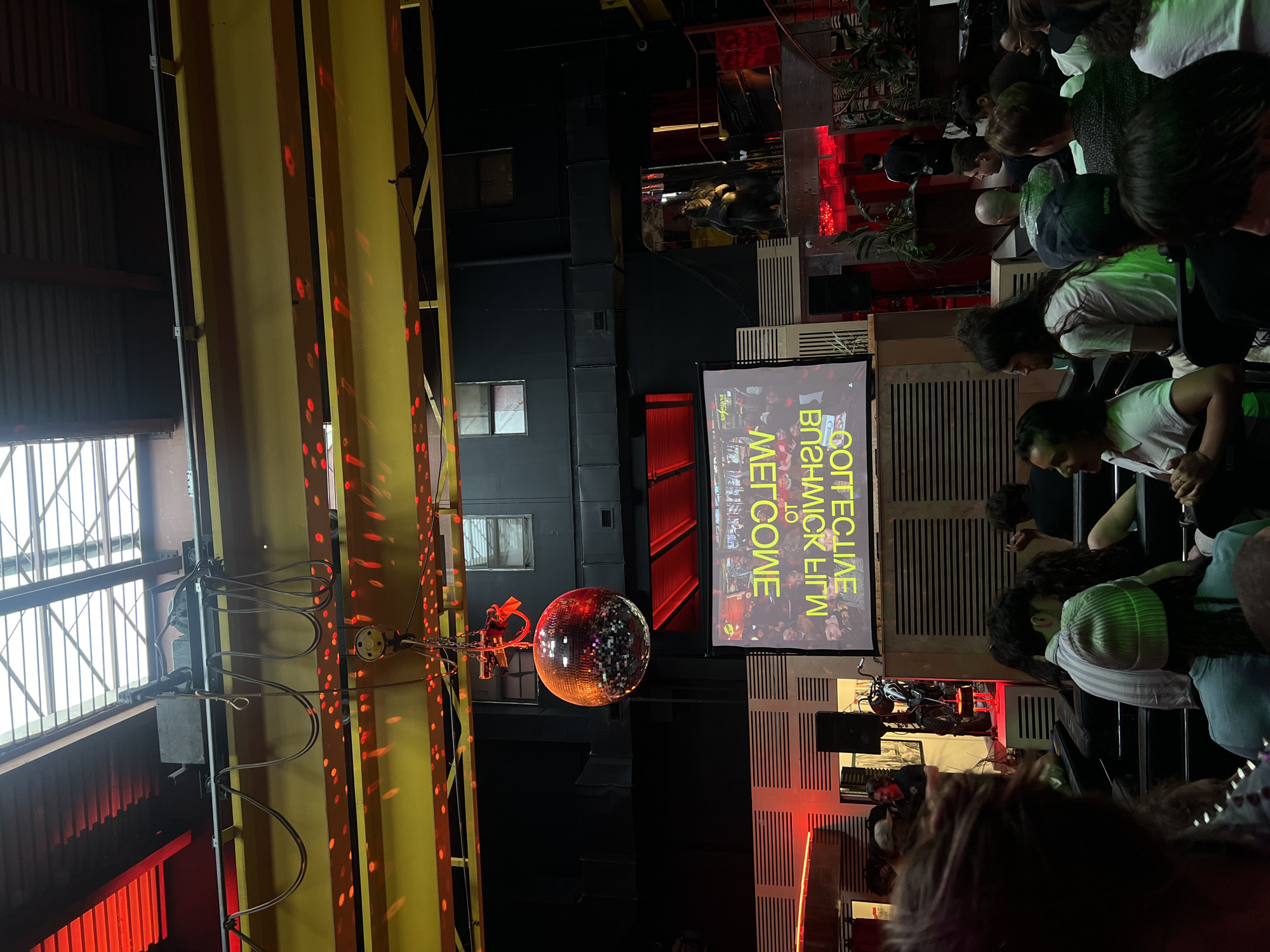 The image shows an indoor event, likely a concert or performance, in what appears to be a club or event space. A disco ball hangs suspended from the ceiling, reflecting light. To the right, there is a screen displaying text, possibly event information. A crowd of people is visible, some standing and watching the event. The lighting is colorful, with red and yellow prominent. The space seems industrial, with exposed ceiling structures and metalwork. The location is not discernible as being in New York City, United States of America from the image.