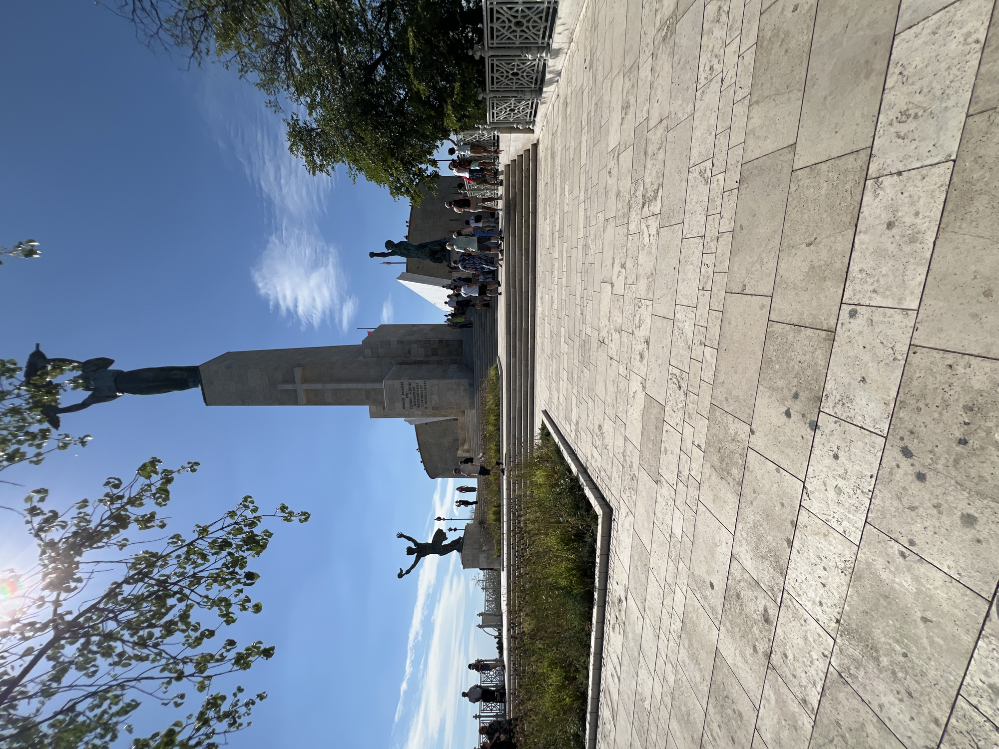 The image depicts the Liberty Monument on Gellért Hill in Budapest, Hungary, on a clear, sunny day. A large, stone monument with a tall, slender obelisk is central to the scene, topped by a statue of a winged female figure holding a palm leaf. A large cross is visible on the front of the obelisk. To the left of the main monument, a second bronze statue depicts a figure with arms outstretched. To the right, a third bronze statue shows a figure with a raised arm.
A wide, paved area with large stone tiles extends from the foreground, leading to a set of wide stairs that ascend towards the monuments. Several people are scattered throughout the scene, some walking up the stairs, others gathered near the statues, and a few standing on a lower level overlooking a landscaped area. Trees are present on the right side of the frame, and branches of a tree with green leaves are visible in the upper left corner, partially obscuring the sun. The sky is a bright blue with scattered white clouds. A decorative metal fence is visible on the right side of the image. The overall impression is of a popular tourist destination with a historical and monumental significance.