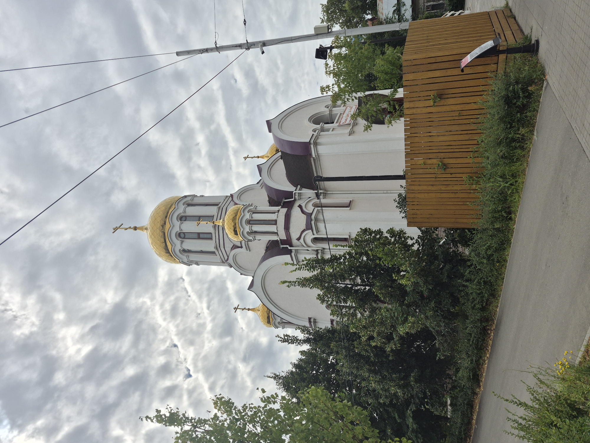 The image shows a white Orthodox church with golden domes, featuring a large central dome and smaller domes on its sides. The church is partially obscured by lush green trees and a tall wooden fence in the foreground. A paved road runs along the left side of the image, bordered by vegetation. Overhead, power lines crisscross the cloudy sky, and a utility pole with attached equipment is visible on the right. In the background, hints of other buildings and more trees suggest an urban or suburban setting. The scene appears to be captured on an overcast day. The church is the Church of the Holy Martyr Elizabeth in Samara, Russia, located in the Soviet district of the city.