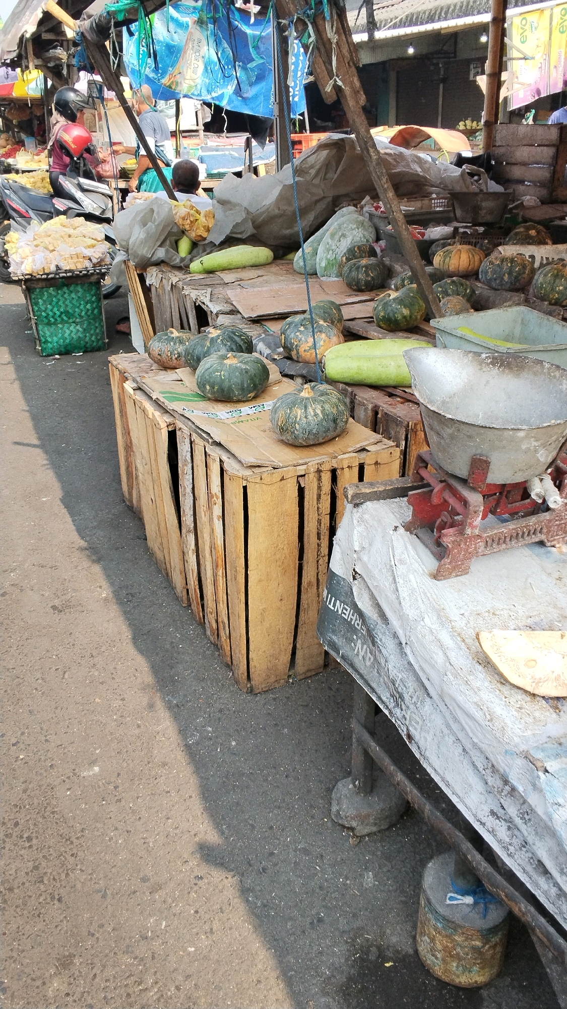 This is an outdoor marketplace scene in Mojokerto, Indonesia. The foreground shows a paved ground with a shadow cast from the left. In the midground, there are several wooden crates, some stacked and others serving as stands. On top of these crates are various vegetables, including pumpkins, gourds, and what appear to be chayotes. Some of the pumpkins are green with orange markings, while others are predominantly green. A large, weathered metal weighing scale with a red base is positioned on a table covered with a grey tarp-like material. In the background, there are market stalls with wooden structures and tarps, displaying more goods and some signage. A few people are visible in the background, including a person in a red helmet sitting on a motorcycle and another person standing near a stall. The overall impression is of a busy, informal market setting.