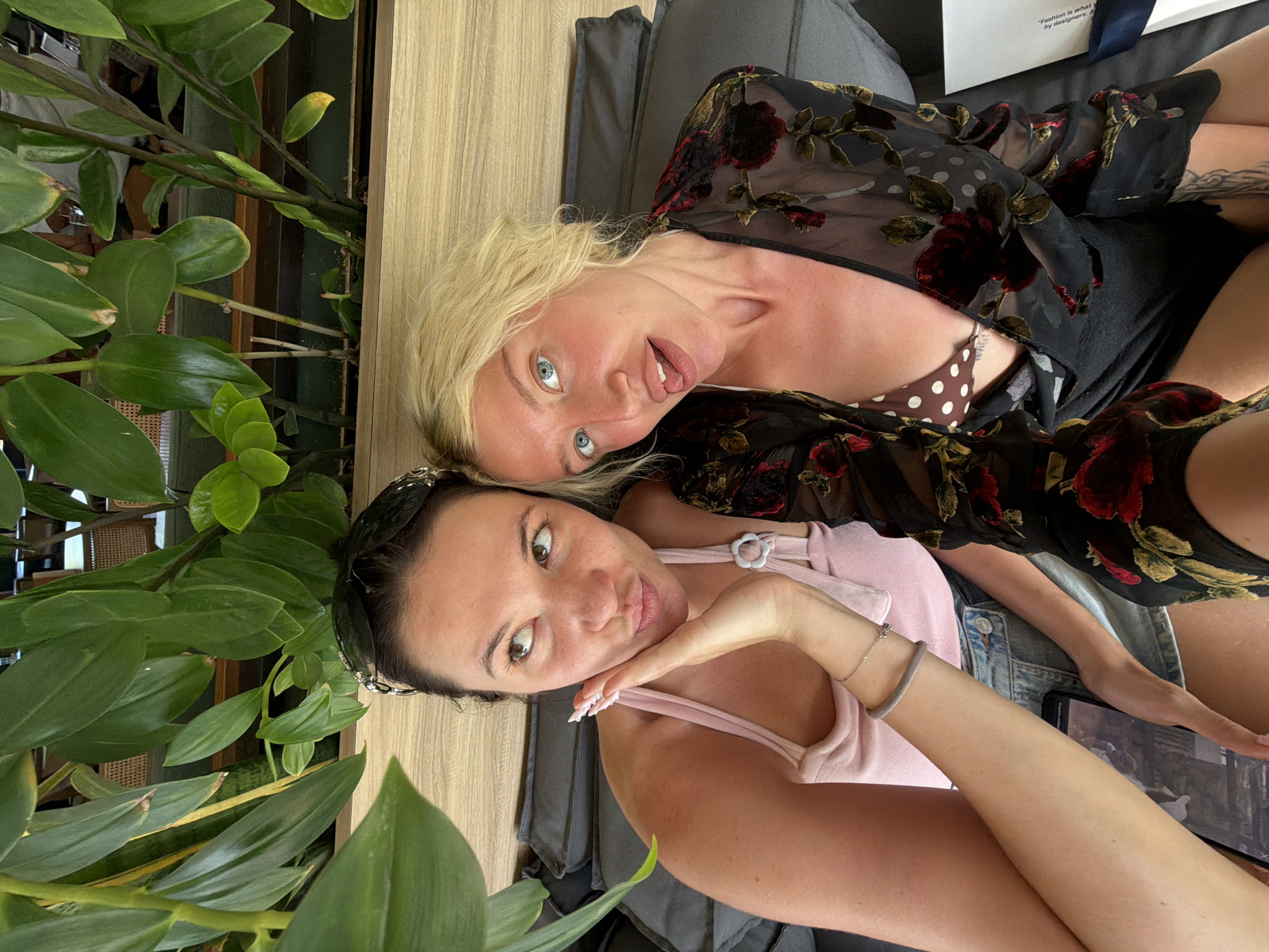 In North Kuta, Indonesia, two women are captured in a selfie taken outdoors, possibly at a cafe or resort, given the lush greenery and casual seating. The woman on the left, with dark hair styled with a headband, wears a pink tank top and denim shorts. She rests her hand on her cheek and puckers her lips, looking upwards. The woman on the right, with blonde hair, is wearing a brown and white polka dot bikini top beneath a sheer black floral kimono. She sticks her tongue out slightly, looking directly at the camera. Both women appear relaxed and are seated on a dark grey cushioned sofa. The background features a variety of plants and a wooden wall.
