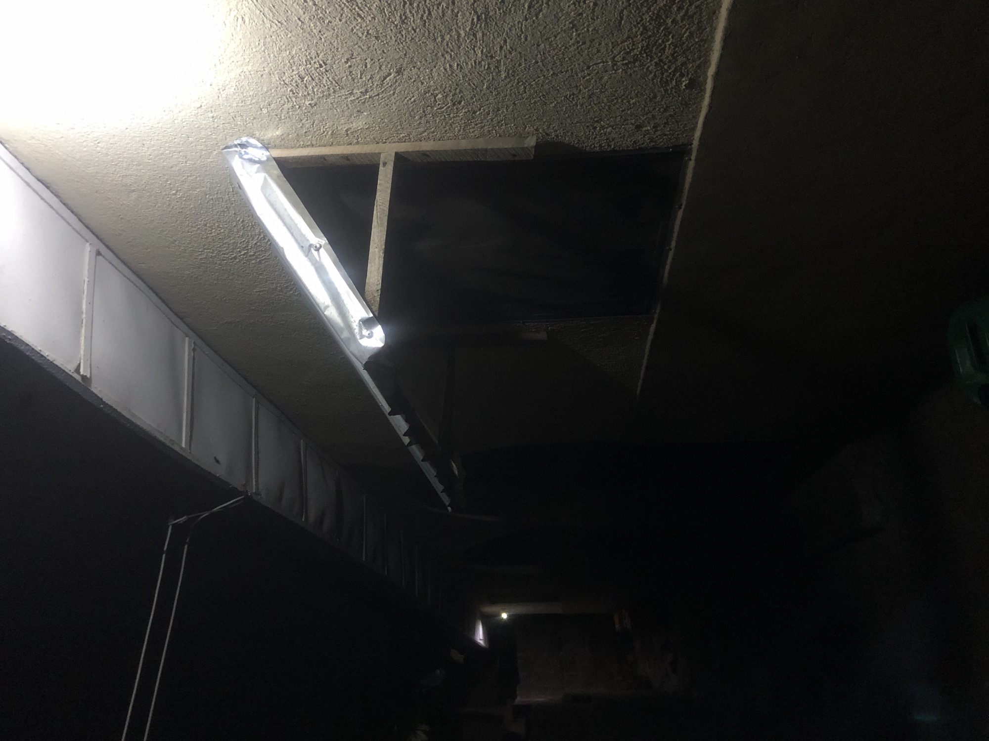 The image captures a dimly lit outdoor scene, likely in Kabong, Nigeria. A fluorescent light fixture is mounted on a textured, yellowish wall, casting a bright glare. Below the light, a dark opening, possibly a window or doorway, is framed by rough wooden planks. To the left, a dark corridor recedes into the distance, with another faint light source visible further down. The ground appears to be unpaved and dark. The overall impression is one of a functional, perhaps utilitarian, outdoor space, illuminated by artificial light in the darkness.