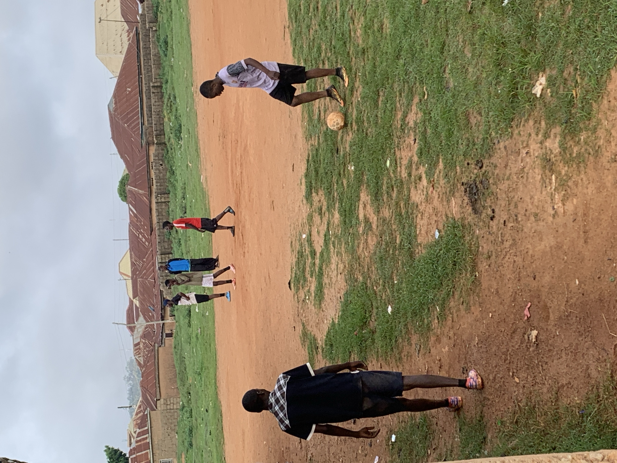 In Bukuru, Nigeria, a group of young men are playing soccer on a dirt field with patches of grass. The sky is overcast, and the surrounding area consists of residential buildings with corrugated iron roofs. One individual in the foreground, viewed from the back, wears a dark short-sleeved shirt with a checkered pattern on the shoulders and dark shorts. Another young man, to the right and facing the viewer, is in mid-play, kicking a soccer ball while wearing a light-colored t-shirt with orange and gray accents and dark shorts. In the background, several other young men are visible, some standing and observing, others potentially participating in the game. The environment appears to be a local community soccer pitch.