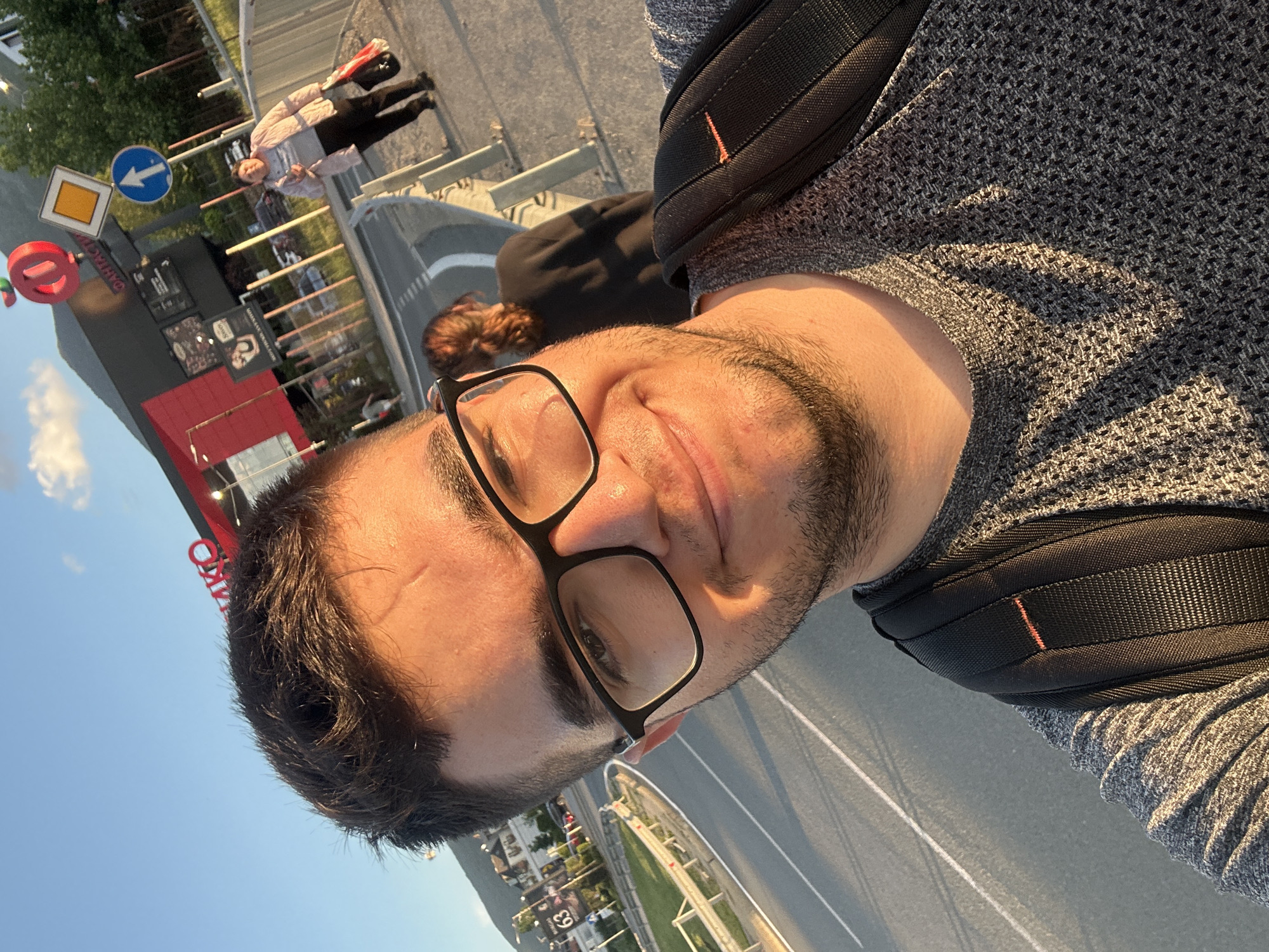 The image is a selfie of a man with dark hair, a beard, and glasses. He is wearing a gray sweater and a black backpack. In the background, there is a street with road markings and a building with a red and black sign. There are also other people visible in the background. The photo appears to have been taken in Sofia, Bulgaria, as indicated by the road signs and general environment. The man is likely standing on a pedestrian bridge or elevated walkway.