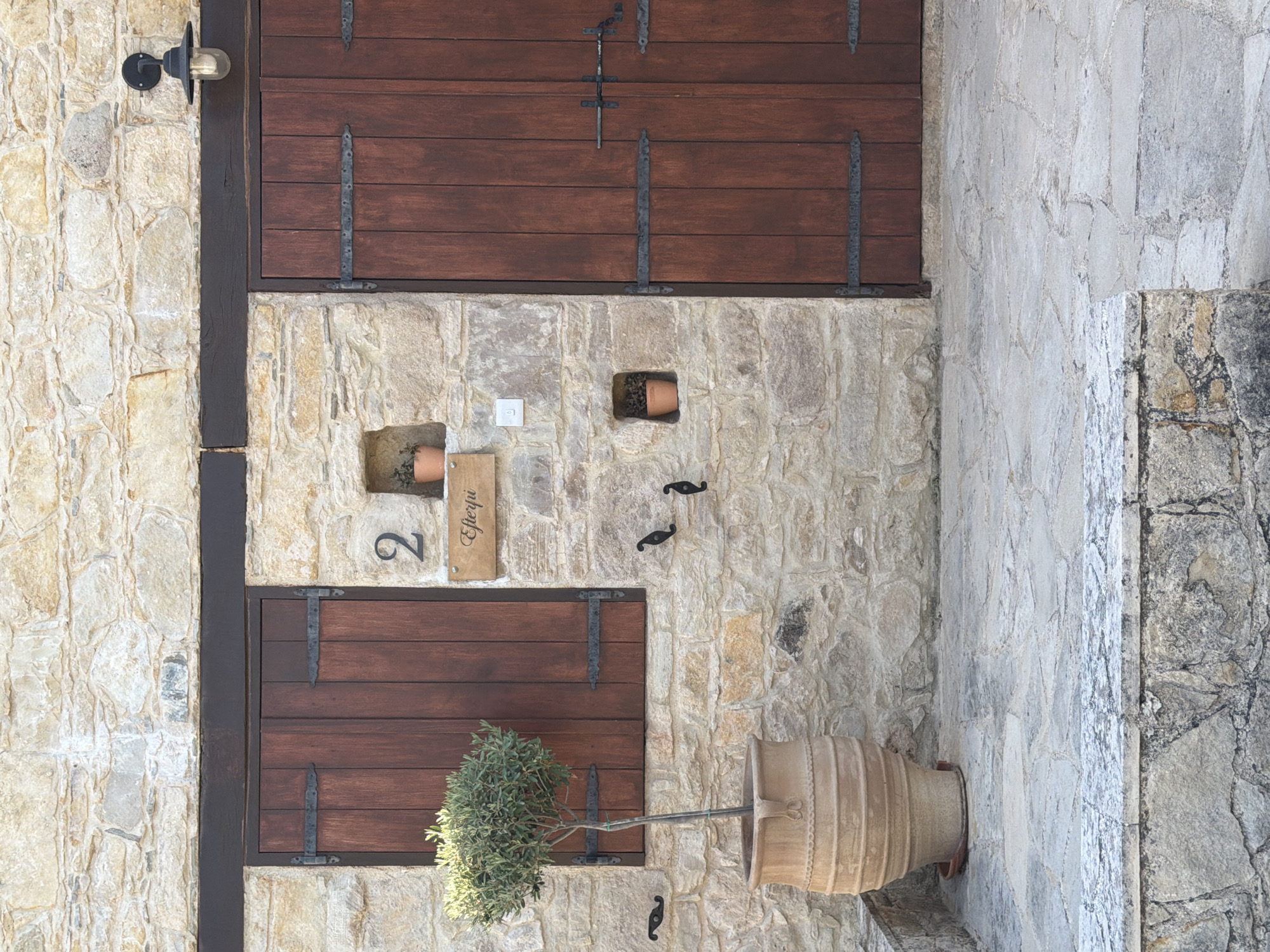 The image shows an exterior view of a stone building with wooden shutters and decorative elements.
The building's facade is constructed from rough-hewn, light-colored stones, giving it a rustic and aged appearance. There are two large, dark brown wooden shutters, one above the other, with visible metal hinges and latches, suggesting a traditional architectural style. To the left of the upper shutter, a black wall-mounted light fixture with a glass cover is visible.
Between the shutters, set into the stone wall, are two small recessed openings. The upper one contains a small terracotta pot with a plant. To the left of this, a small, light-colored square object, possibly an electrical outlet cover, is mounted on the stone. Below the upper shutter and to the right of the lower shutter, there are two small, black, decorative metal elements attached to the wall, resembling elongated S-shapes.
Below the upper shutter, to the left, there is a recessed terracotta pot with a plant. To its right, a light-colored wooden sign is mounted, with black cursive text that appears to read "Efterpi". Next to the sign, a black numeral "2" is affixed to the stone.
The lower shutter is adorned with similar dark metal hinges and latches. In front of the lower shutter, a small, leafy green plant is growing from a pot that is not fully visible. To the right of the lower shutter and extending across the frame, a large, light-colored terracotta amphora-like pot is positioned on the ground, containing a small, dense green tree. Another black, decorative metal element is mounted on the stone wall below the lower shutter.
The scene appears to be outdoors, with a cobblestone or paved pathway visible on the right side of the image, made of large, irregularly shaped stones. The lighting suggests it is daytime, with a soft, even illumination, indicating possibly overcast or late afternoon light. The overall impression is one of a well-maintained, charming property with a Mediterranean or rustic aesthetic.