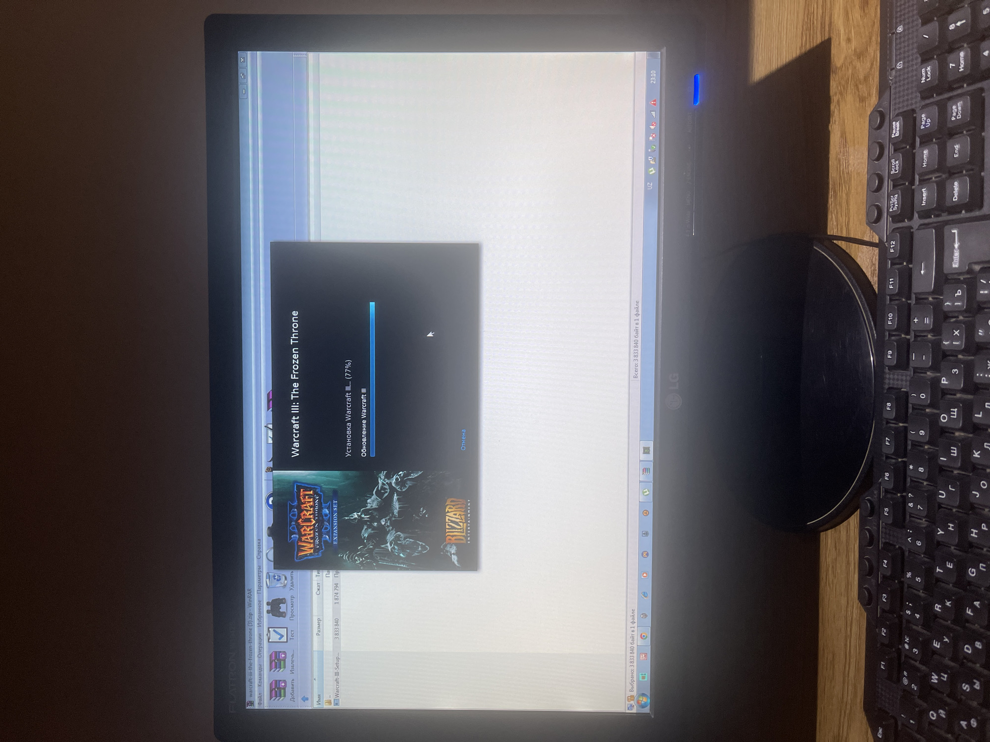 The image shows a computer monitor displaying an installation progress window for "Warcraft III: The Frozen Throne". The progress bar indicates the installation is at 77%. In the background, a file explorer window from WinRAR is visible, showing a file named "Warcraft-III-the-frozen-throne (3).zip". The monitor is placed on a wooden surface, likely a desk, and a portion of a black keyboard is visible in the foreground. The overall scene suggests a user is in the process of installing or updating the game. There is no visible indication of the location being in Qarshi, Uzbekistan.
