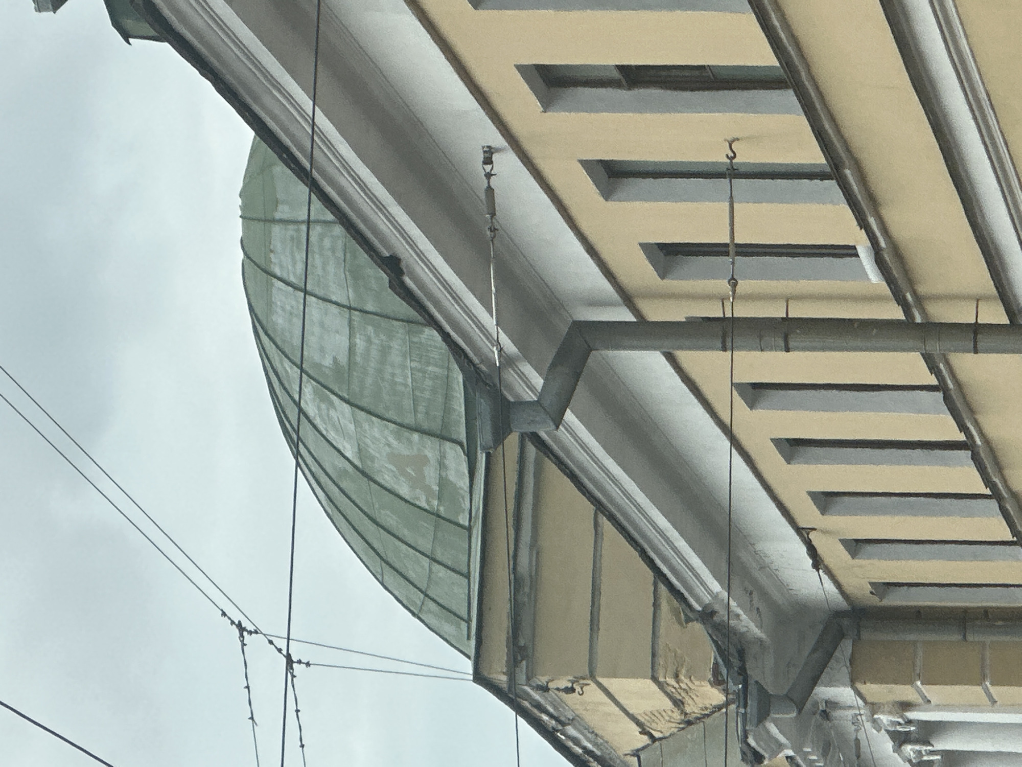 The image depicts a close-up, ground-level perspective of the facade of a historic building in Saint Petersburg, Russia. The building features a light yellow exterior with a white ornate cornice. Above the cornice, the upper part of a green, segmented dome is visible, characteristic of many architectural landmarks in the city. Overhead, a network of electrical wires crisscrosses the frame, suggesting the presence of a tram or trolleybus line, common in the urban landscape of Saint Petersburg. A downspout made of metal is affixed to the side of the building, directing water away from the structure. Several windows with dark frames are aligned along the facade, spaced evenly apart. The sky is overcast and grey. The image appears to have been taken on a cloudy day.