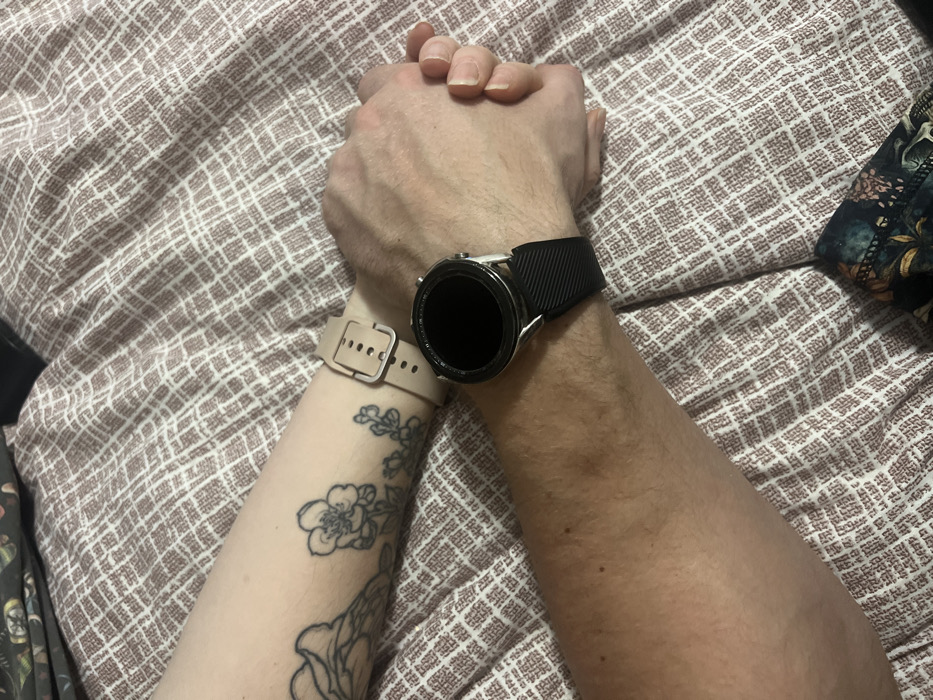 Two arms on a textured beige and brown surface hold hands, with one arm showing a black tattoo of flowers and a smartwatch with a pale peach band. The other arm wears a black smartwatch. The skin tone varies noticeably between the two arms, with one appearing significantly darker. A patterned fabric is visible in the corners.