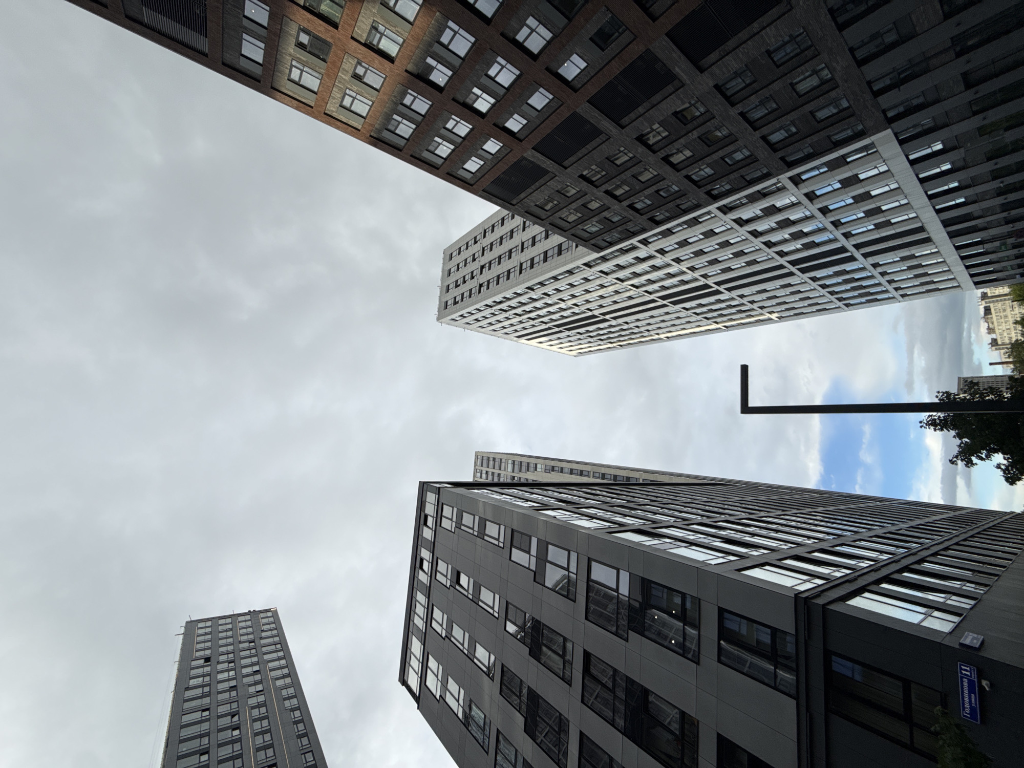 The image shows a low-angle view of several tall buildings against an overcast sky. The buildings have a modern architectural style with many windows. One building is brown and black brick, another is white with black detailing, and another is gray with dark window frames. A streetlight is visible. A street sign in Russian reads "Годовикова 11" (Godovikova 11), indicating the location is Godovikova Street in Moscow, Russia.