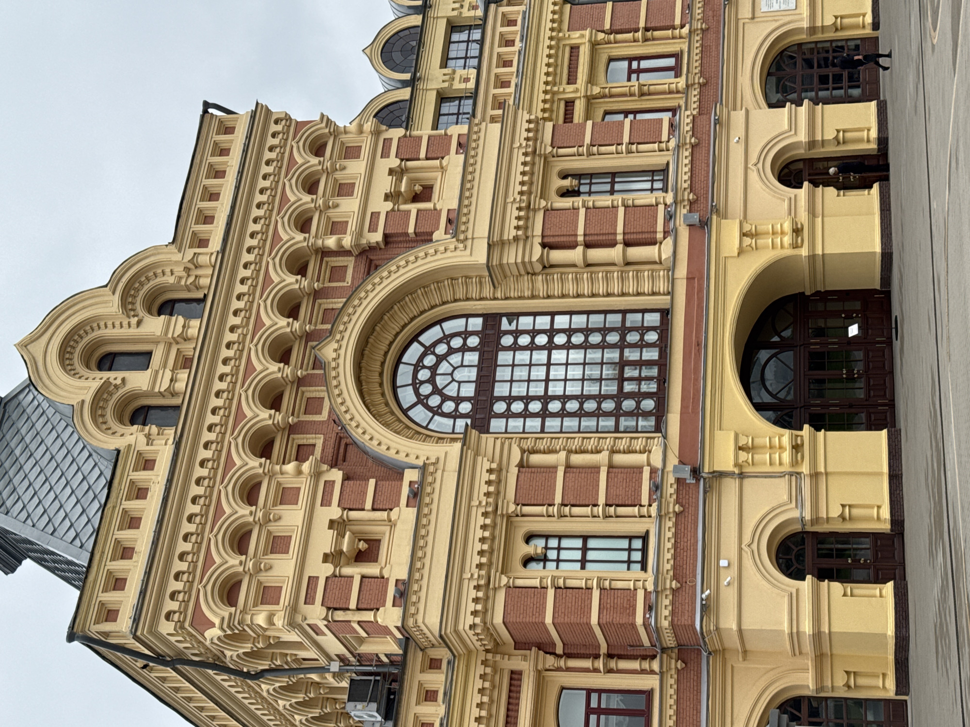 The image shows a detailed view of the facade of the Nizhny Novgorod State Bank building, located on Bolshaya Pokrovskaya Street in Nizhny Novgorod, Russia. The building features a striking combination of red brick and yellow stucco, with intricate architectural details. Prominently displayed is a large, arched window with a grid pattern, flanked by smaller windows. The lower portion of the building showcases arched doorways and decorative columns. Two individuals are visible in the foreground; one is a person in dark clothing walking away from the camera, and another is standing near an entrance. The sky is overcast, indicating a cloudy day.