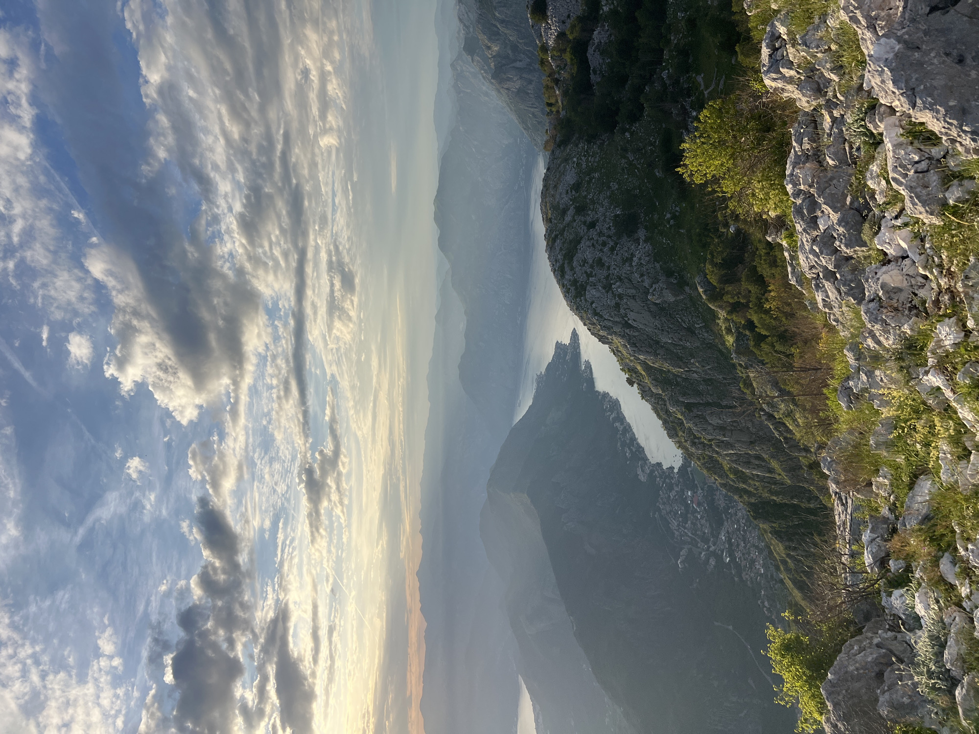 This is a high-angle, panoramic landscape photograph taken from an elevated rocky vantage point in Kuk, Montenegro, overlooking a large body of water that winds through mountainous terrain. The scene depicts a late afternoon or early morning setting, indicated by the soft, warm light and atmospheric haze.
The upper third of the image displays a partly cloudy sky, with patches of clear blue interspersed among scattered cumulus and stratus clouds of varying densities, ranging from bright white to light grey. Several thin, white linear contrails are visible in the upper sky.
Below the sky, multiple mountain ranges extend into the distance, exhibiting increasing atmospheric perspective, which renders the farthest peaks in soft blue-grey tones. A significant, serpentine body of water, characteristic of a bay or fjord, dominates the mid-ground, reflecting the sky's light and appearing calm. Along the shores of this water body, particularly on the left, are visible clusters of buildings forming small, conglomerate settlements nestled at the base and on the lower slopes of the mountains.
The immediate foreground consists of rugged, light-toned rocky terrain. Scattered among these rocks are sparse patches of green vegetation, including grasses, small bushes, and low-lying shrubs. No human figures or specific actions are depicted within the frame. The primary subjects are the natural geological formations, the water body, and the distant built environment.