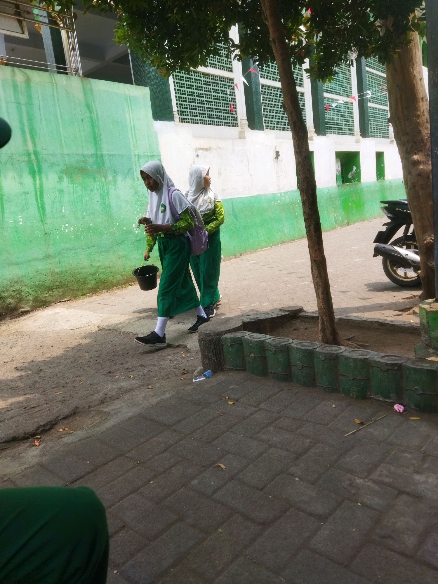 Two young females in green hijabs and long green skirts walk on a paved walkway. The female in the foreground is carrying a black bucket in her right hand and wearing a white top with green trim on her sleeves. The female behind her is wearing a white hijab and a green top, and is carrying a purple backpack. They are walking next to a bright green wall with streaks of white paint and a white building with green window accents. Trees are present in the foreground and background. A dark motorcycle is parked on the right side of the image. The ground is paved with interlocking bricks and dirt. There are some fallen leaves and a pink object on the ground. The scene appears to be an outdoor area near a school or religious institution, likely in Pasrepan, Indonesia, given the typical attire and architecture.