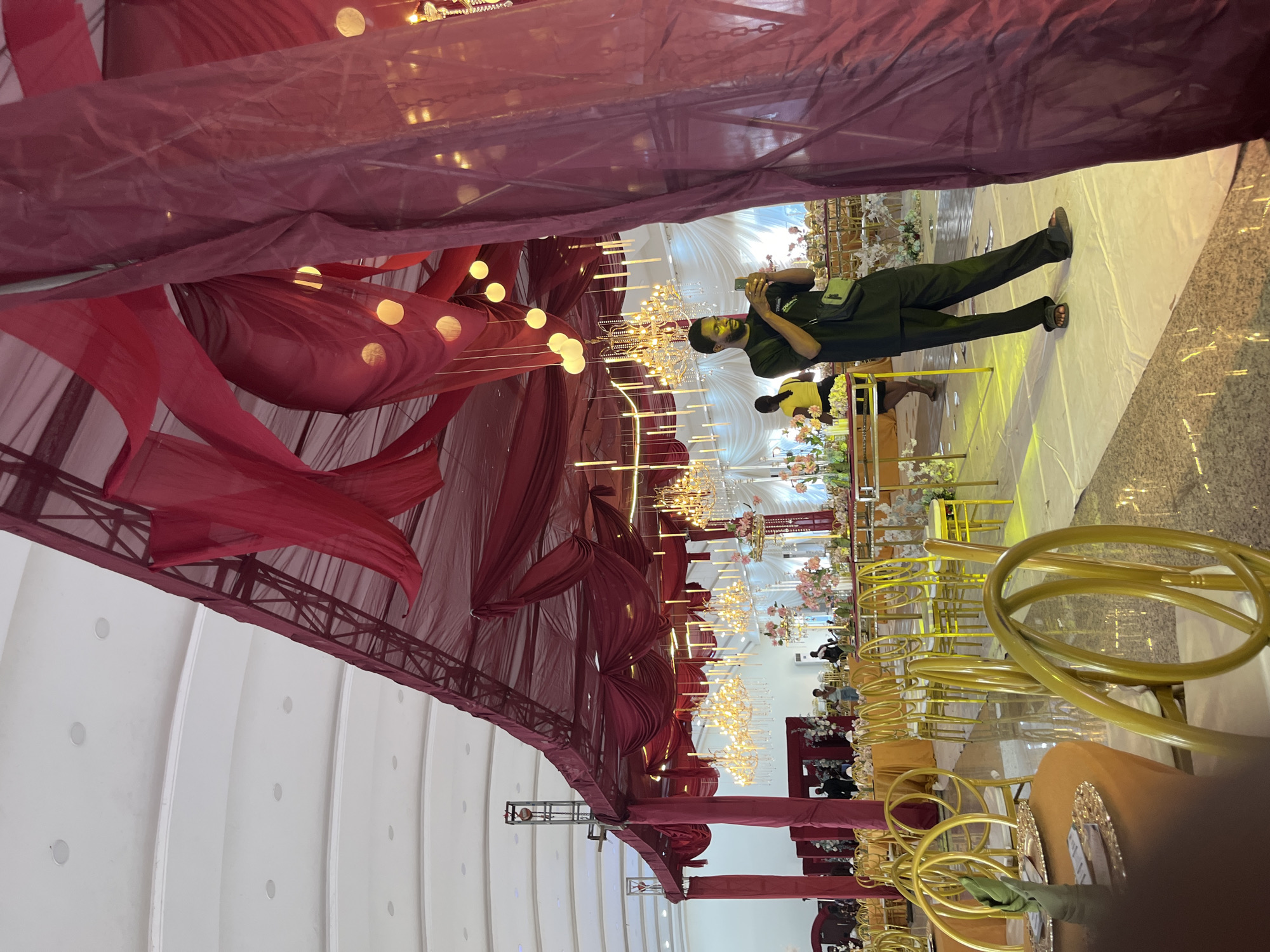 The image depicts a decorated indoor event space, possibly a ballroom or reception hall, featuring a burgundy draped ceiling with hanging lights and chandeliers. In the foreground, a man in a black shirt and pants is standing and taking a photo with his phone, his body angled slightly to the right. He is wearing a cross-body bag. Behind him, a woman in a yellow top and black skirt is visible, walking away from the camera. The space is set up with numerous round tables and gold chairs, some of which are in focus in the foreground. Floral arrangements are placed on tables. The lighting suggests it is daytime, with natural light entering from the ceiling. The overall scene is one of a formal gathering or celebration.
